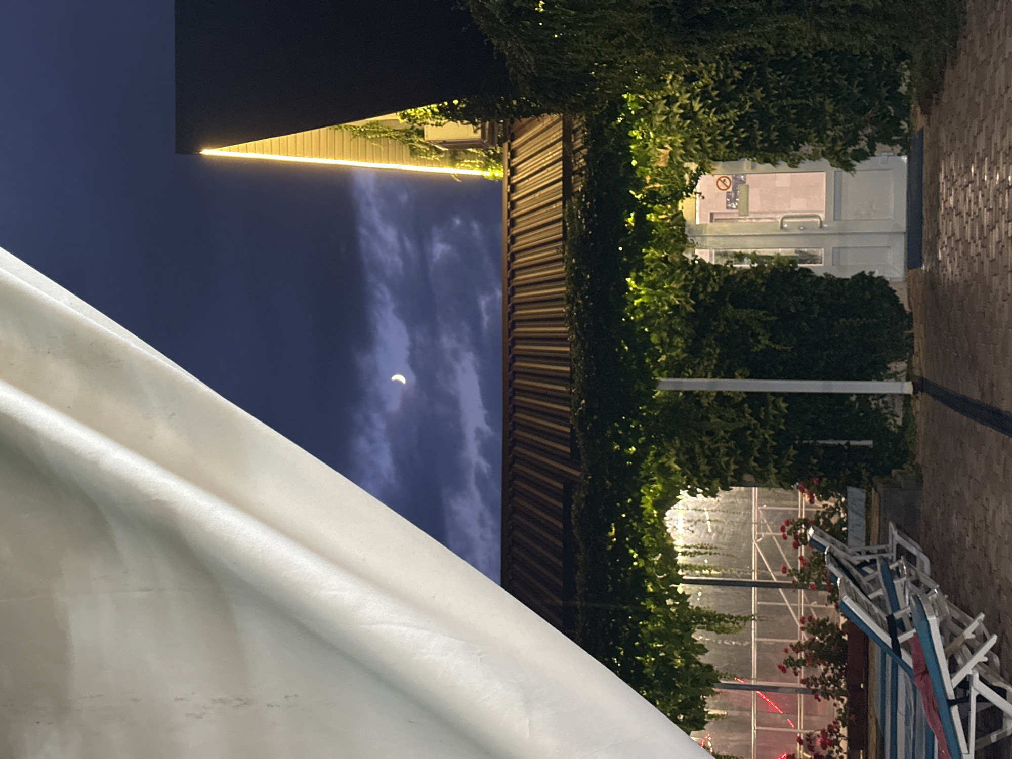 The image depicts a nighttime scene in Yaroslavskaya, Russia, likely an outdoor seating area or resort. In the foreground, a large, white canvas drapes across the left side of the frame, partially obscuring the view. Below the canvas, a corrugated metal roof is visible, covered in lush green vines. A pathway paved with interlocking bricks leads towards a white door framed by more dense green foliage. The door has a silver handle and a frosted glass panel with some signage visible. Red flowers in planters line the area to the left of the pathway, alongside several white lounge chairs with blue and white striped cushions. In the upper portion of the image, the night sky is a deep blue, with scattered clouds and a crescent moon. A modern building with a dark facade and illuminated yellow lighting along its edge is visible on the right. The overall impression is one of a tranquil, dimly lit outdoor space at dusk or night.
