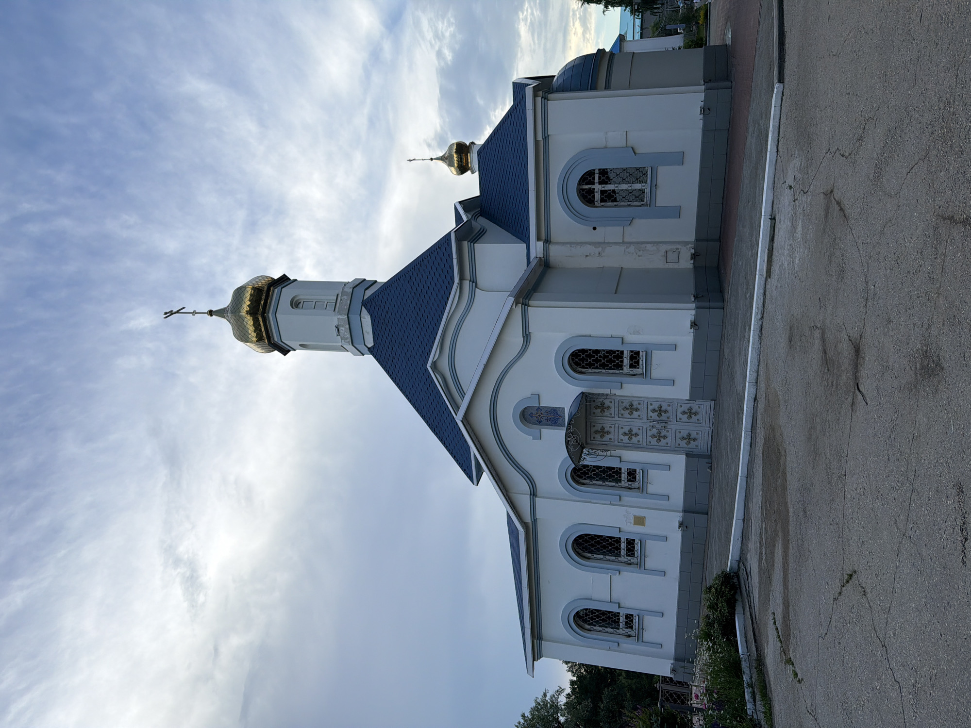 An Eastern Orthodox church is pictured under a cloudy sky. The church is white with a blue roof and a golden dome topped with a cross. It has several arched windows with decorative grilles and a large, ornate double door at the front. The building is situated on a paved area with some cracked asphalt in the foreground and a small patch of grass and flowers to the left. Trees are visible behind the church to the left and on the right. The church is located in Volzhskiy, Russia.