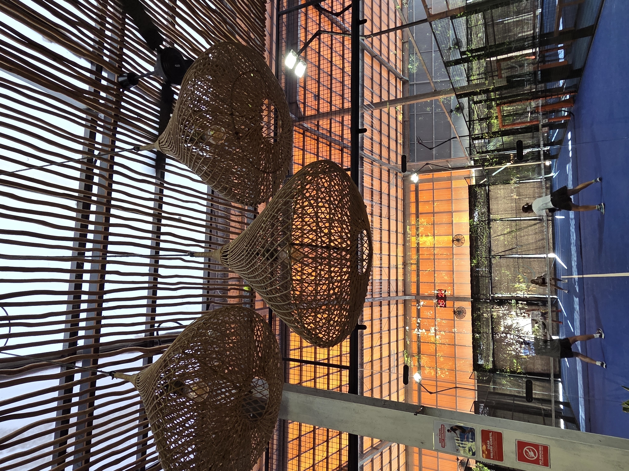 This media file depicts an indoor padel tennis court located in North Kuta, Indonesia. The setting features a blue court with white lines, enclosed by a metal fence and a roof. The lighting suggests it is either late afternoon or evening, with artificial lights illuminating the court. Sunlight filters through a slatted wooden ceiling and an orange-tinted glass structure, casting warm hues.
Several people are visible on the court, engaged in playing padel. One individual, wearing a white t-shirt and black shorts, is in motion near the net. Another person in a dark shirt and black shorts is also visible on the court. In the background, other players and spectators can be seen.
Hanging from the ceiling are three large, woven basket-like pendant lamps, adding a decorative and rustic element to the indoor space. The overall atmosphere appears to be active and recreational.
Visible text includes a sign that reads "MINI PADEL ONLY BOOKING" and another sign with an image and text, possibly a warning or information about playing. A "NO SMOKING" symbol is also visible on a red sign. The time displayed on a digital clock on the wall reads "16:25".