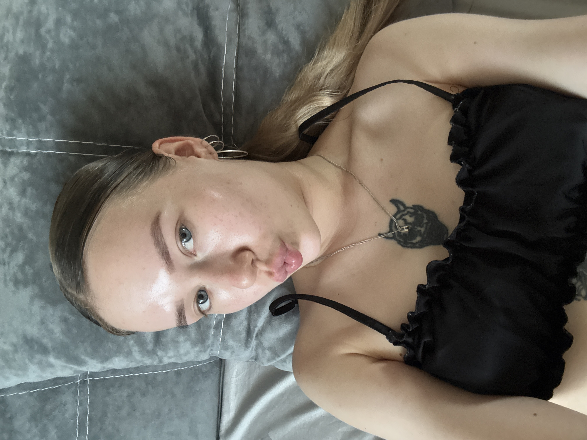 A young woman lies on a gray, quilted surface, possibly a bed or cushion. She has fair skin, light blue eyes, and her hair is blonde, pulled back sleekly at the front and flowing loose towards her right. She's wearing a black top with ruffled edges and thin straps. A delicate silver necklace rests on her chest, with a black tattoo visible below it. She's puckering her lips, creating a "kiss" expression. There is no evidence to confirm the location is Chaikovsky, Russia.