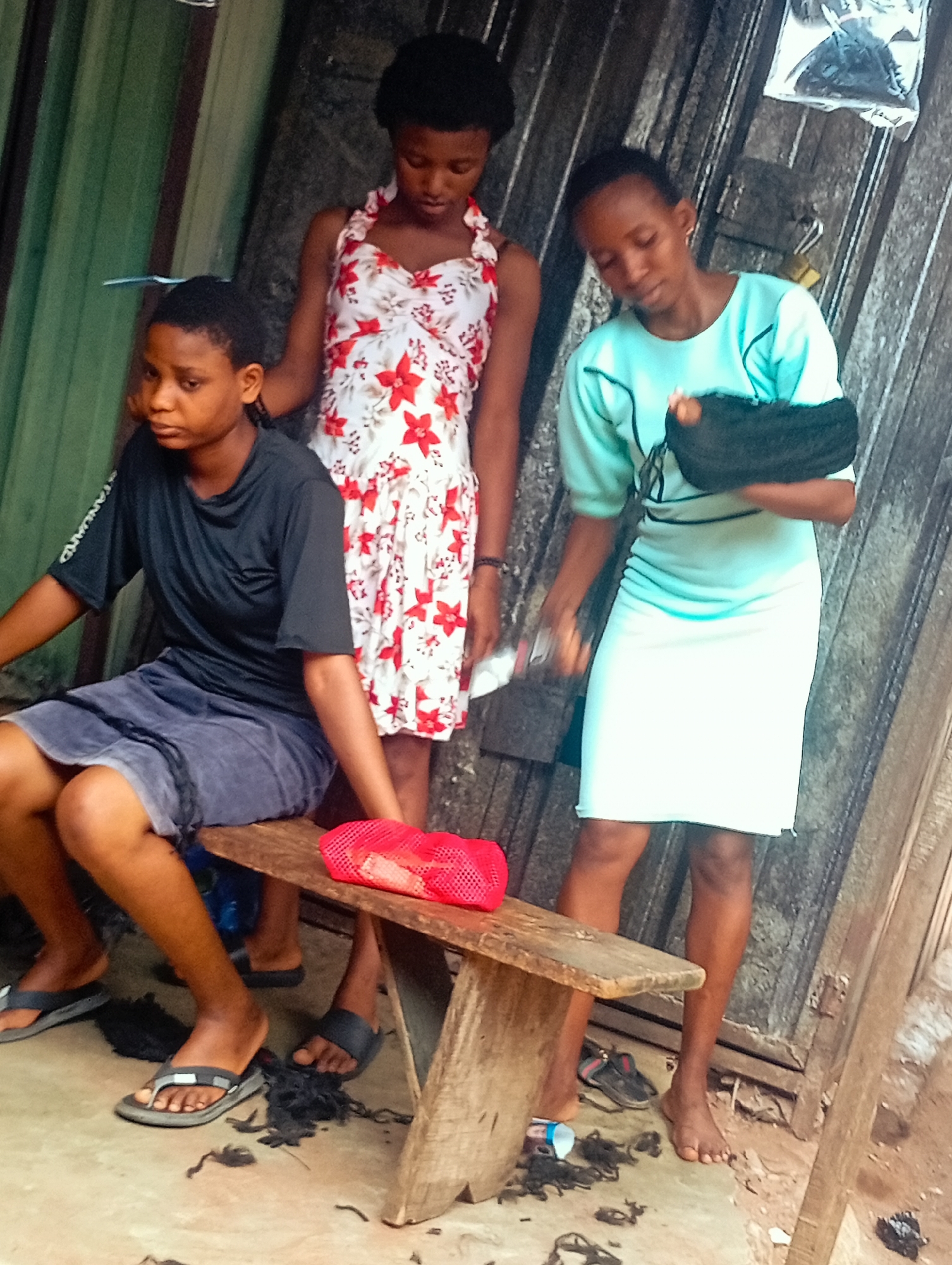 Three young people are positioned in an outdoor setting, likely a shop or stall with a corrugated metal backdrop. A young person on the left is seated on a wooden bench, wearing a black t-shirt and gray shorts. They have a neutral expression and their hair is cut short. To their right stands another young person in a white dress with red floral patterns. They are looking down, holding a white object in their right hand. On the far right, a third young person stands wearing a light blue top and a white skirt. Their left arm is in a black sling, and they are looking downwards.
The ground around them is dirt and scattered with cut hair, indicating a haircut is in progress or has recently concluded. A red mesh bag sits on the bench, and a pair of black flip-flops are visible on the ground near the seated person. Another pair of black sandals is near the feet of the person with the sling. A plastic bottle and more cut hair are scattered beneath the bench. The lighting suggests it is daytime, possibly mid-morning or afternoon, with diffused light. The overall atmosphere appears casual and practical, with the focus on the hair cutting activity. There is no visible text in the image.