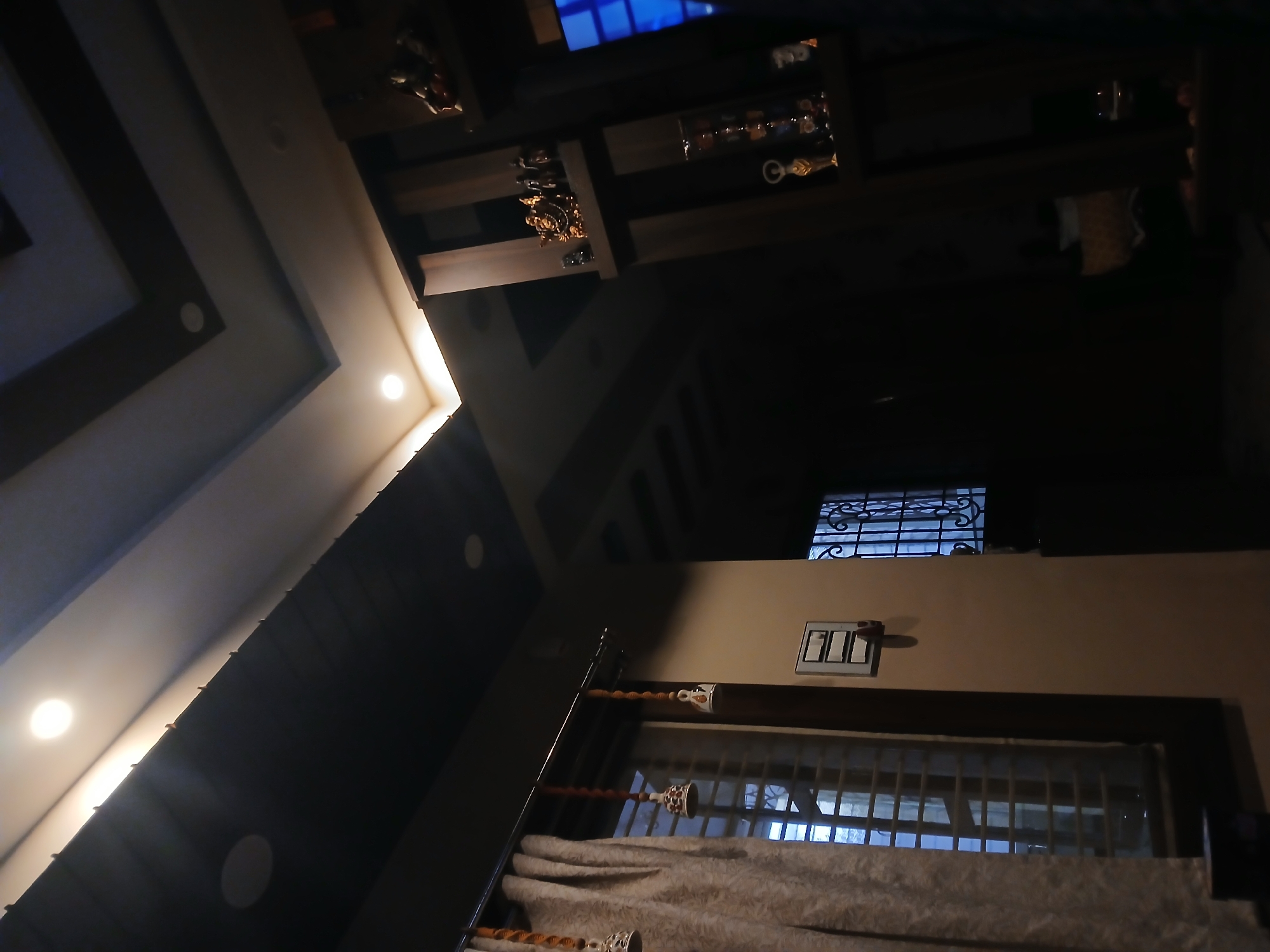 A low-angle interior shot, likely in Bengaluru, India, depicts a room with a mix of modern and traditional elements. The ceiling on the left is a multi-tiered design with recessed lighting and brown accents. Above, there is what seems to be a display case with decorative objects, possibly figurines or glassware. On the right, a wall features a window with white curtains and a decorative wrought-iron grill above it. A light switch is also visible on the wall. The lighting is dim, contributing to the contrast between the lit ceiling and the darker areas of the room. The general impression is that of a well-furnished living space that is dimly lit.