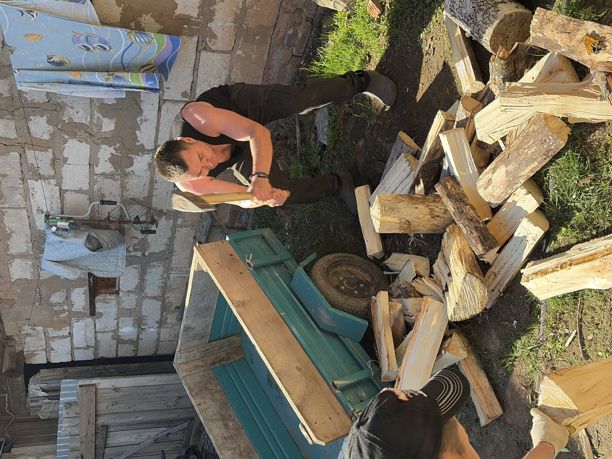 The image shows a man chopping wood in what appears to be a backyard setting, possibly in Nizhnii Vishur, Russia. He is dressed in dark clothes and boots, wielding an axe over a pile of logs. A small, green trailer is nearby, partly loaded with firewood. The background includes a cinder block wall and some hanging linens, suggesting a domestic setting. The area is partially covered in grass and dirt, indicating an outdoor environment. Another person is visible near the wood pile.