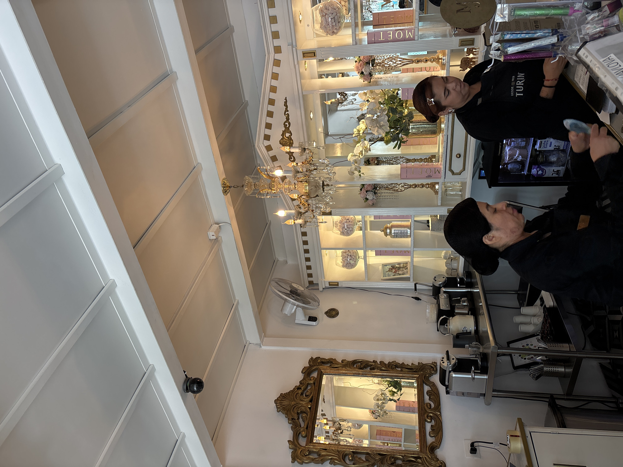 Two female employees stand behind a counter in what appears to be a confectionery or dessert shop in San Bernardino Tlaxcalancingo, Mexico. One woman is holding a small, light-blue object in her hand. Behind the counter are shelves displaying decorative items and confectionery products, including what appears to be Moët champagne advertisements. A chandelier hangs from the ceiling above. A decorative gold-framed mirror is on the wall and reflecting the shop's interior. A fan is mounted on the wall above the mirror. Multiple coffee machines and accessories are visible behind the counter. The scene has an elegant and somewhat ornate aesthetic.
