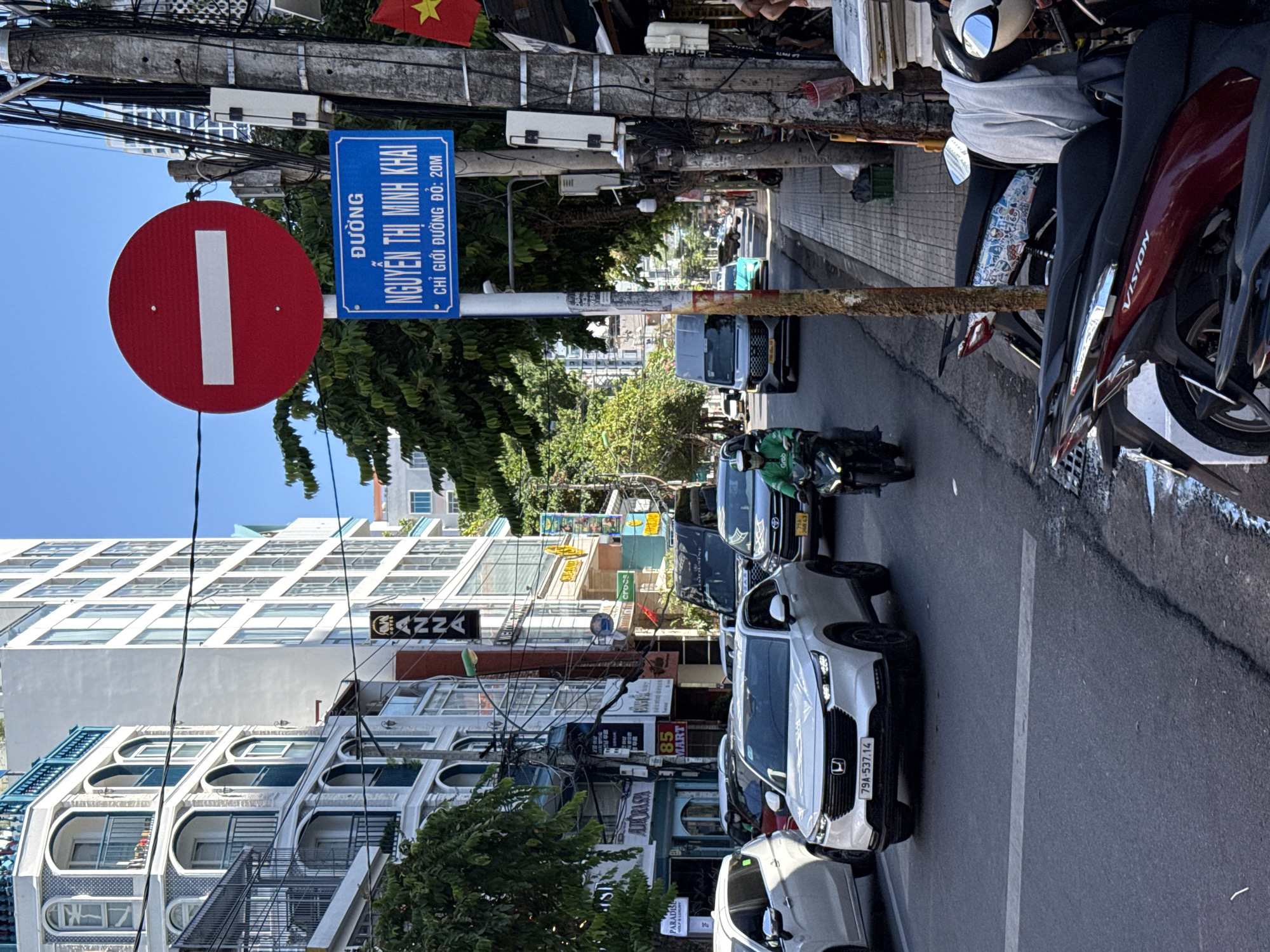 In Nha Trang, Vietnam, a street scene unfolds under a clear blue sky. A white SUV, license plate 79A-537.14, is parked on the right side of the road. A motorcyclist wearing a green jacket rides a black motorcycle, also heading right. To the left, a silver van is visible. Further down the street, buildings of varying heights line the road. A blue street sign, reading "ĐƯỜNG NGUYỄN THỊ MINH KHAI" with the text "CHỈ GIỚI ĐƯỜNG ĐÔ: 20M" below, is mounted on a pole. A circular red sign with a white horizontal bar, indicating "Do Not Enter," is positioned above the street sign. The sidewalk on the right is lined with parked motorcycles and some stalls or shops. Wires and utility poles are prominent features of the streetscape.
