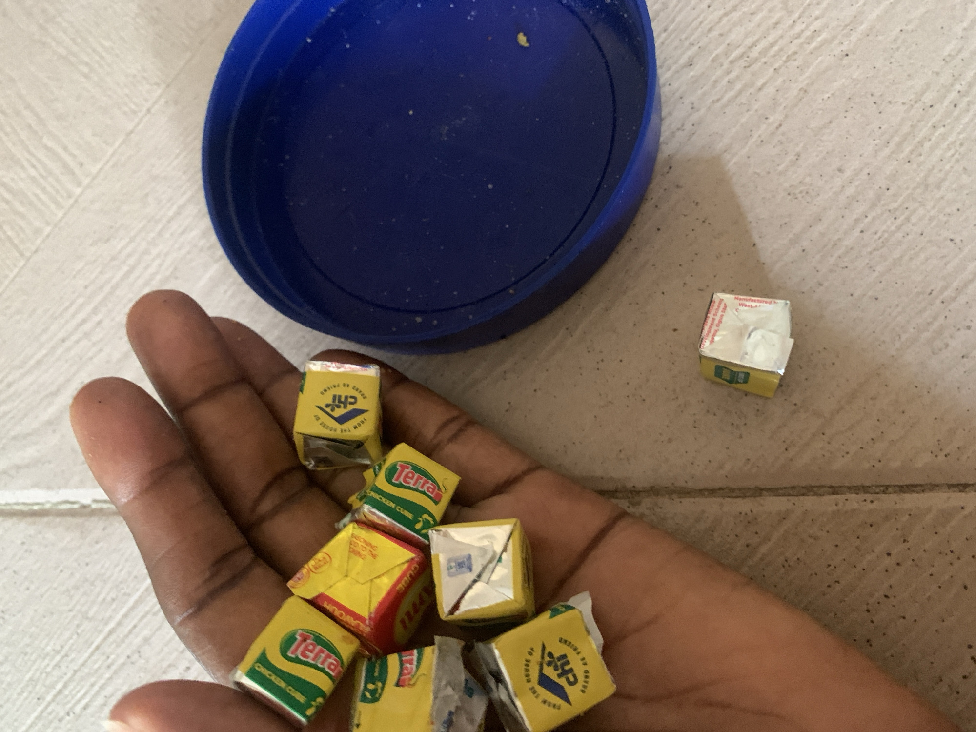 A person's hand, holding several yellow bouillon cubes, rests on a tiled surface. A blue plastic lid is situated to the right of the hand. One bouillon cube lies on the floor near the lid. The cubes appear to be branded with "Terra" and "Chi" logos.