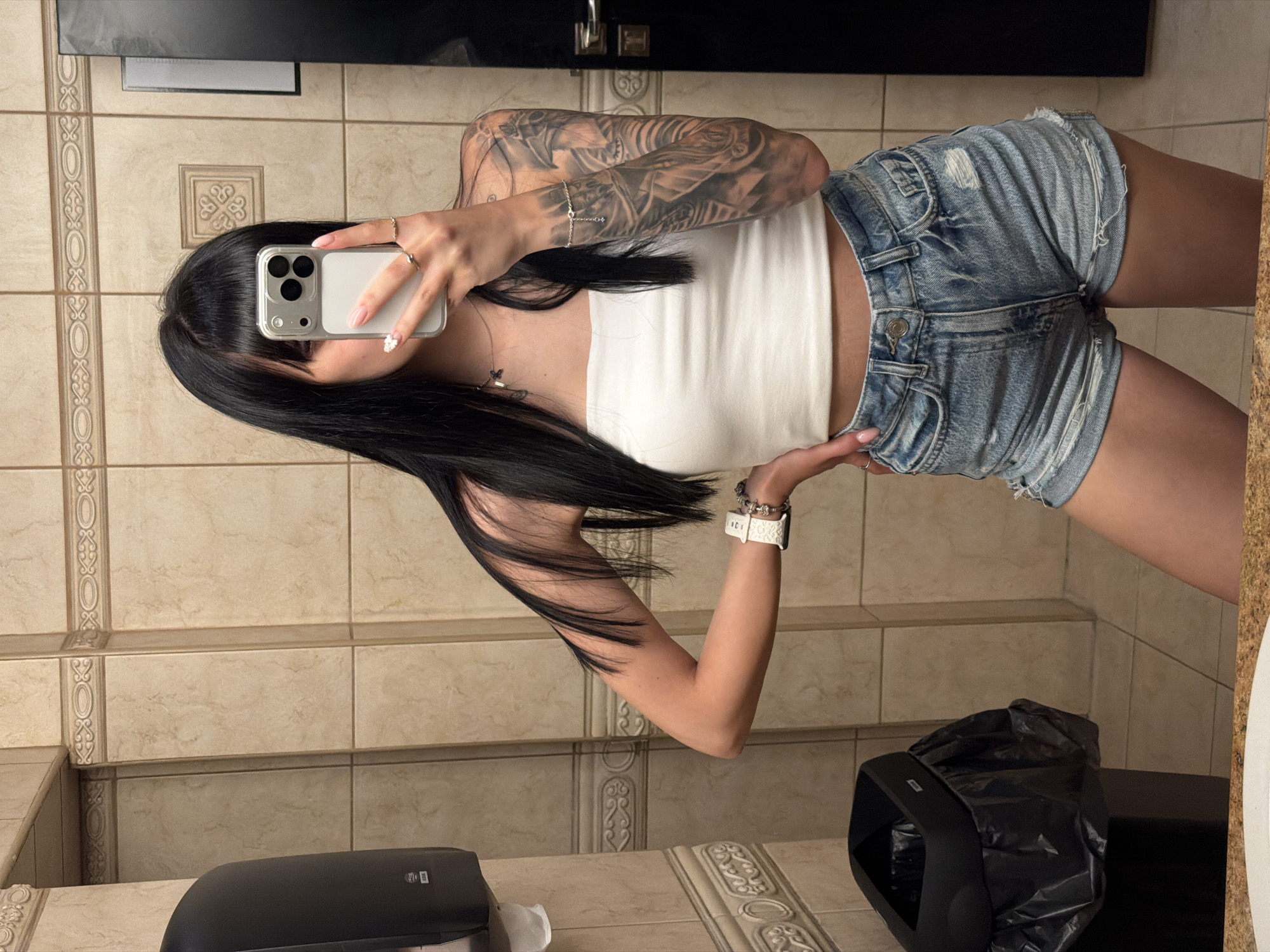A female individual is captured in a mirror selfie within what appears to be a public restroom. She has long, dark, straight hair with bangs, which partially obscures her face as she holds a light-colored smartphone with a triple camera array, covering her upper facial features. Her right hand, adorned with rings and light-colored nails (one featuring a small white floral detail), holds the phone. She is wearing a white strapless crop top and light blue distressed denim shorts. Her left arm is covered by a prominent, dark-toned tattoo sleeve depicting intricate patterns and possibly figures, and she also wears a white wristwatch and additional bracelets on this arm. A delicate silver-colored necklace with a small charm is visible around her neck. Her left hand rests on her hip.
The background consists of light beige/tan rectangular tiled walls. Two horizontal decorative tile borders with scrollwork and oval patterns traverse the wall at different heights, and a single square decorative tile is visible above the subject's head. To the left, a black wall-mounted hand dryer and a black trash can with a liner are present. The bottom edge of a light-colored, speckled sink counter is visible in the foreground. To the right, a section of a dark-colored door or stall door with a silver handle and lock mechanism is visible. The specific location in the city of Plovdiv, Bulgaria, cannot be determined from the visual information provided in the image.