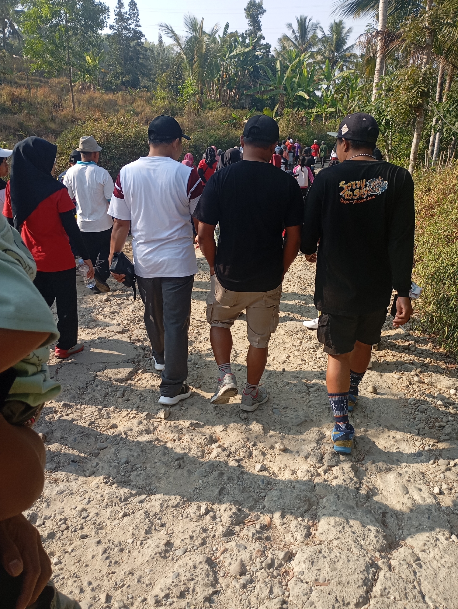 The image depicts a group of people walking on a dirt road in a rural area, likely Ketepung, Indonesia, given the presence of lush vegetation including palm trees and banana trees, which are common in the region. The road is unpaved and appears to be made of dirt, gravel, and small stones, with evidence of erosion in some areas. The sunlight casts distinct shadows on the ground, suggesting it is daytime.
In the foreground and midground, several individuals are seen from the back, walking away from the camera. One person in a black long-sleeved shirt with a graphic print on the back is wearing dark shorts and patterned socks with athletic shoes. To their left, another person in a black t-shirt and khaki shorts is also walking away. Next to them, a person in a white t-shirt with maroon trim and grey pants is walking, holding a black bag. Further to the left, a person wearing a red shirt and black pants, with a black headscarf, is visible. A person in a white t-shirt and a grey hat is also present. In the background, more people are seen walking further down the road, interspersed with dense green foliage and tall palm trees. The overall scene suggests an outdoor activity or journey in a tropical, possibly agricultural, setting.