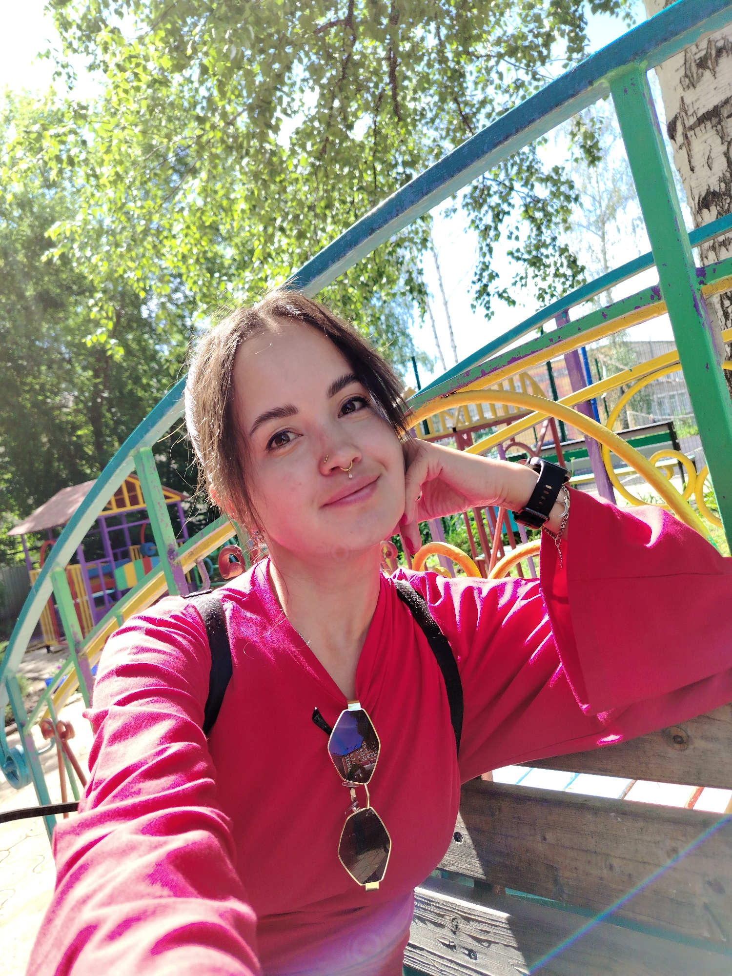 A young woman with dark hair, fair skin, and a nose ring is taking a selfie outdoors. She is wearing a bright pink, loose-fitting shirt with bell sleeves and a black backpack. Sunglasses with gold frames are hanging from the neckline of her shirt. She is resting her head on her hand, and her left arm, adorned with a black smartwatch and bracelets, is bent. The background features a colorful children's playground structure with blue, green, yellow, and purple elements, as well as trees and a wooden bench. The lighting suggests a sunny day. The presence of a birch tree on the right side of the image is consistent with the landscape of Chaikovsky, Russia.