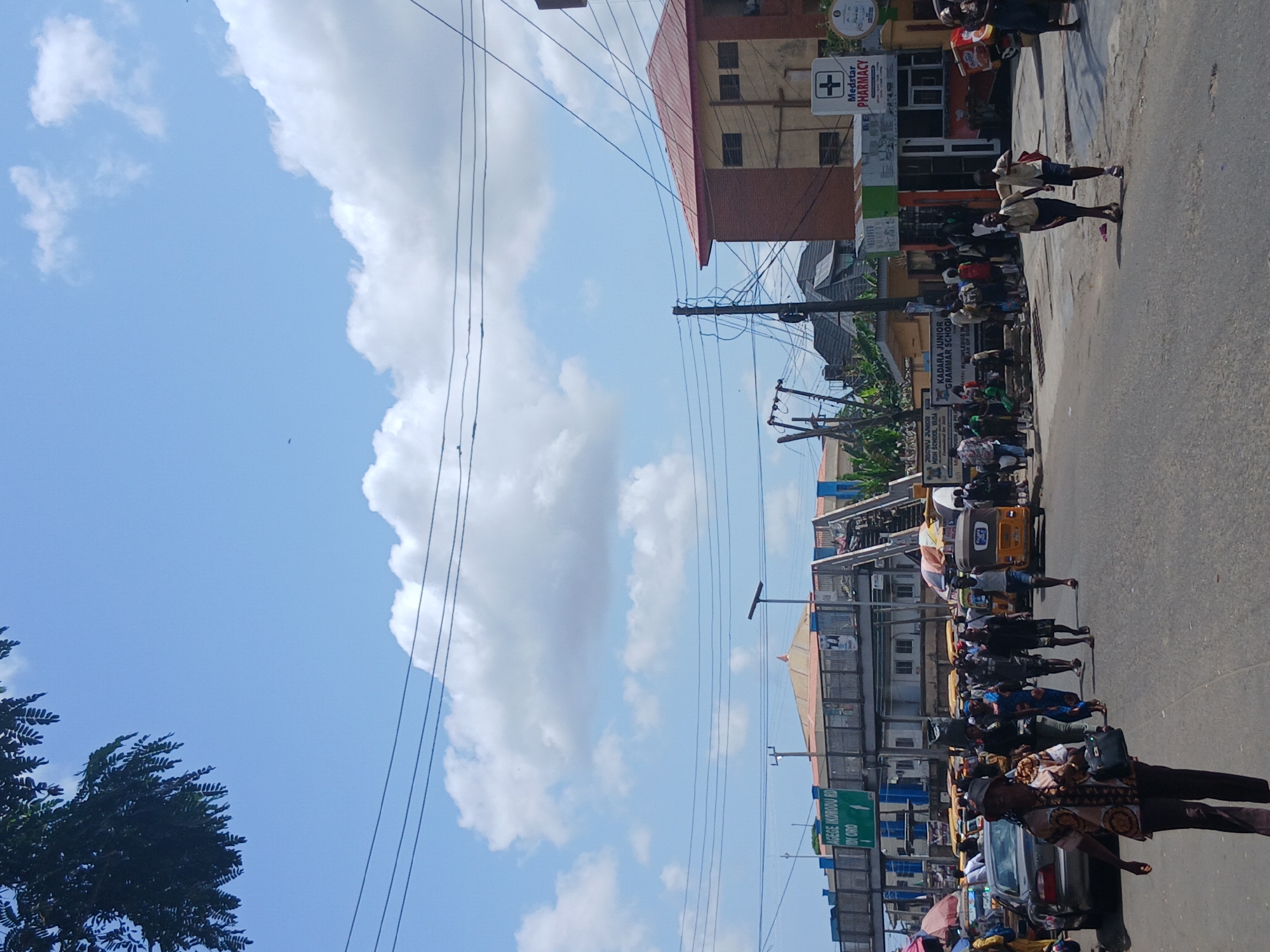 The image shows a street scene in Lagos, Nigeria, on a sunny day with scattered clouds. The view is along a road bustling with pedestrians, some of whom are carrying bags or luggage. A number of yellow "Keke Napep" (autorickshaws) are visible in the middle of the road. Several buildings line the street, including what appears to be a pharmacy (Medstar Pharmacy) and a school (Kadara Junior Grammar School). A street sign for "Isege Korodu Rd Idi Oro" is also visible. Power lines crisscross the sky.