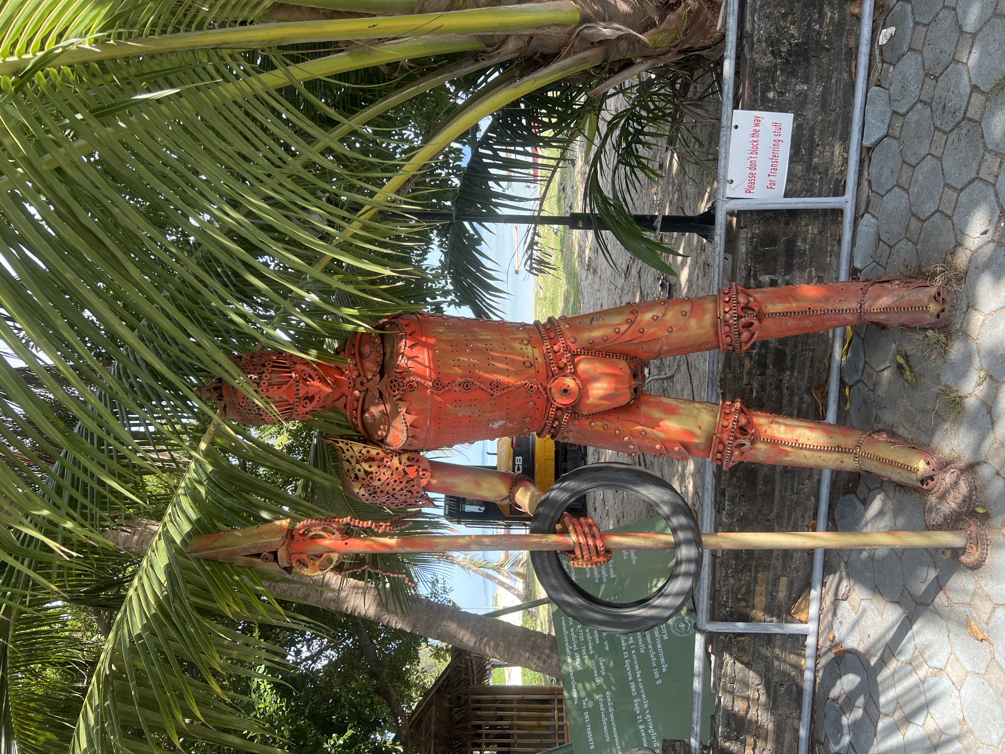 A full-body shot captures a statue of a knight made of rusted, orange-painted metal, standing beneath lush green palm trees. The knight is positioned centrally, facing forward, and holds a spear with a tire attached to it in its right hand. The statue's armor is intricate, with visible chains and gears. Behind the statue, a sandy beach with clear blue water and a few boats can be seen. A yellow construction vehicle, possibly a small excavator, is parked on the beach. A small wooden shack with a thatched roof is visible to the left of the statue. A sign with Thai writing is displayed on a green board in the background, and a white sign reading "Please don't block the way For Transferring stuff" is attached to a metal railing in the foreground, to the right of the statue. The ground in front of the statue is paved with hexagonal stone tiles, and scattered fallen leaves are visible. The image appears to be taken on a sunny day, with shadows cast by the palm trees and the statue on the ground. The location is identified as Ko Phangan, Thailand.