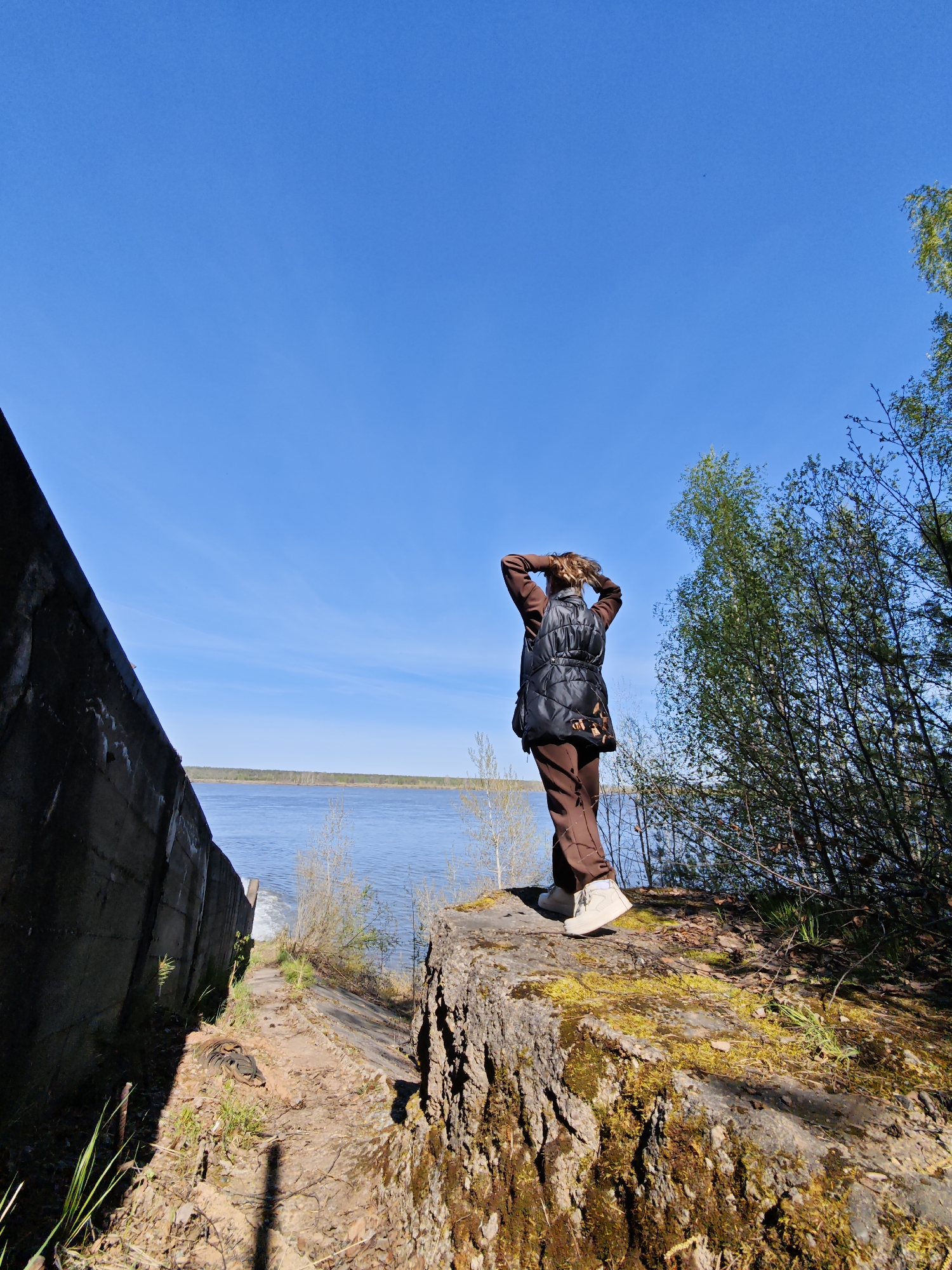 A woman stands on a moss-covered stone structure overlooking a body of water. She wears brown pants, a dark vest, and white shoes. Her hands are behind her head. The background includes a concrete wall, trees, and a clear blue sky. The location is Chernushka, Russia.