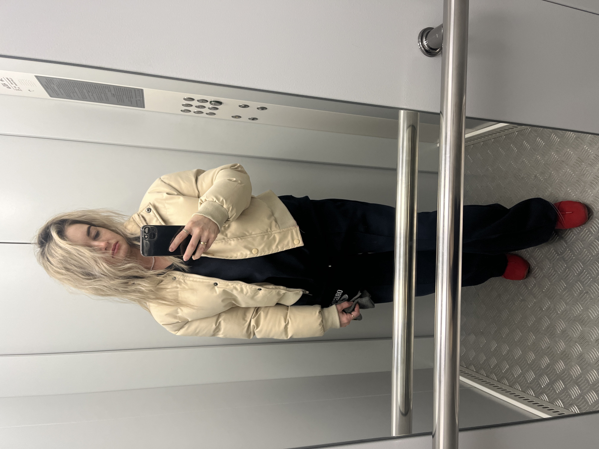 A female is standing in an elevator taking a picture of herself. She is wearing a light-colored puffer jacket, black pants, and red shoes. She is holding a black smartphone in her right hand. The elevator has a stainless-steel interior with a control panel and handrails. The floor is made of textured metal. The photo was taken in the city of Chaikovsky, Russia.