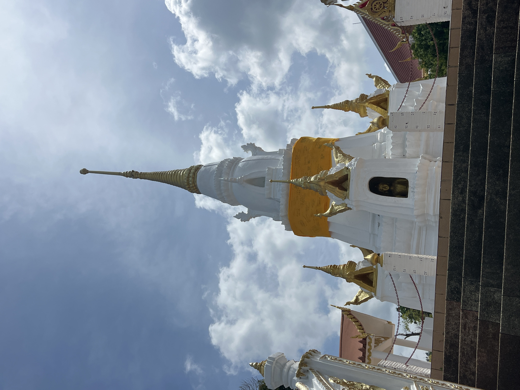 A low-angle shot captures a white stupa with golden spires and accents against a partly cloudy sky. The stupa is decorated with a bright yellow sash around its midsection. A small golden Buddha statue is visible in a niche on the stupa's lower level. Several smaller golden spires adorn the sides of the main structure. In the foreground, a set of dark stone stairs leads up towards the stupa. The background shows hints of red-tiled roofs of other buildings and lush green trees. The scene is set at Wat Phra Yai, also known as the Big Buddha temple, located in Baan Hua Thanon, Thailand, a well-known landmark on Koh Samui island.