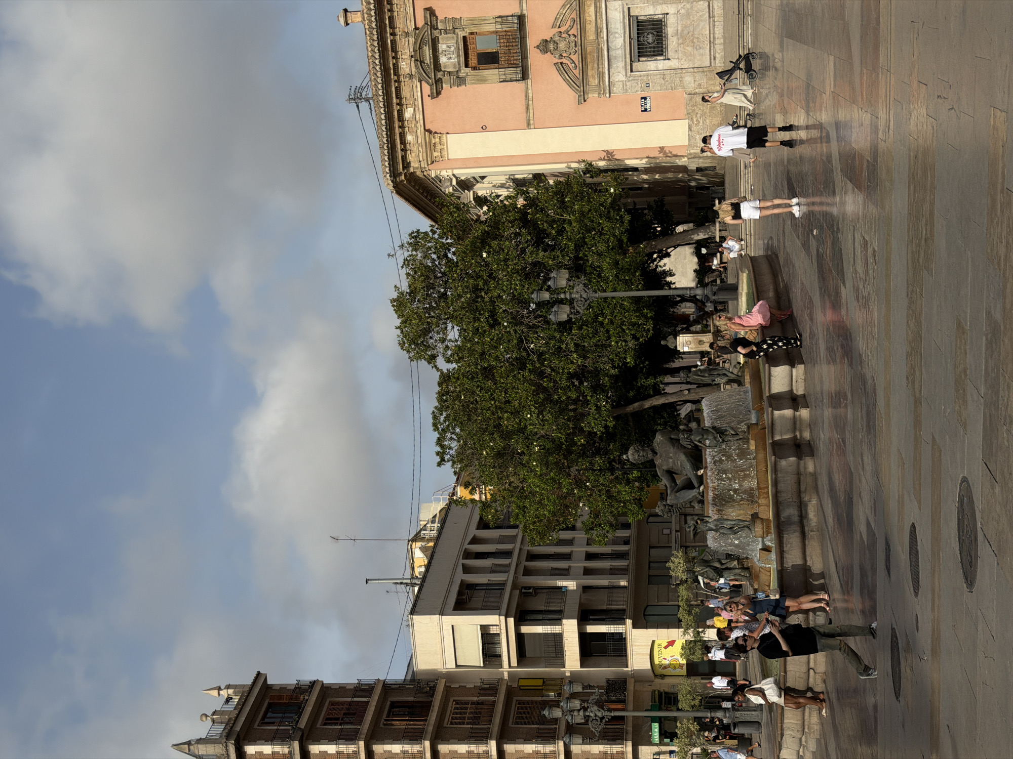 The image depicts a European-style plaza with a large fountain as the central element. Several people are present in the plaza, some interacting with the fountain, while others are walking or standing. The architecture surrounding the plaza includes a multi-story building with balconies and a larger, ornate pink building with a distinctive window. A large tree provides shade in the vicinity of the fountain. The ground is paved with stone tiles. The sky is partly cloudy. A sign on the pink building reads "PLAZA DE LA VIRGEN".