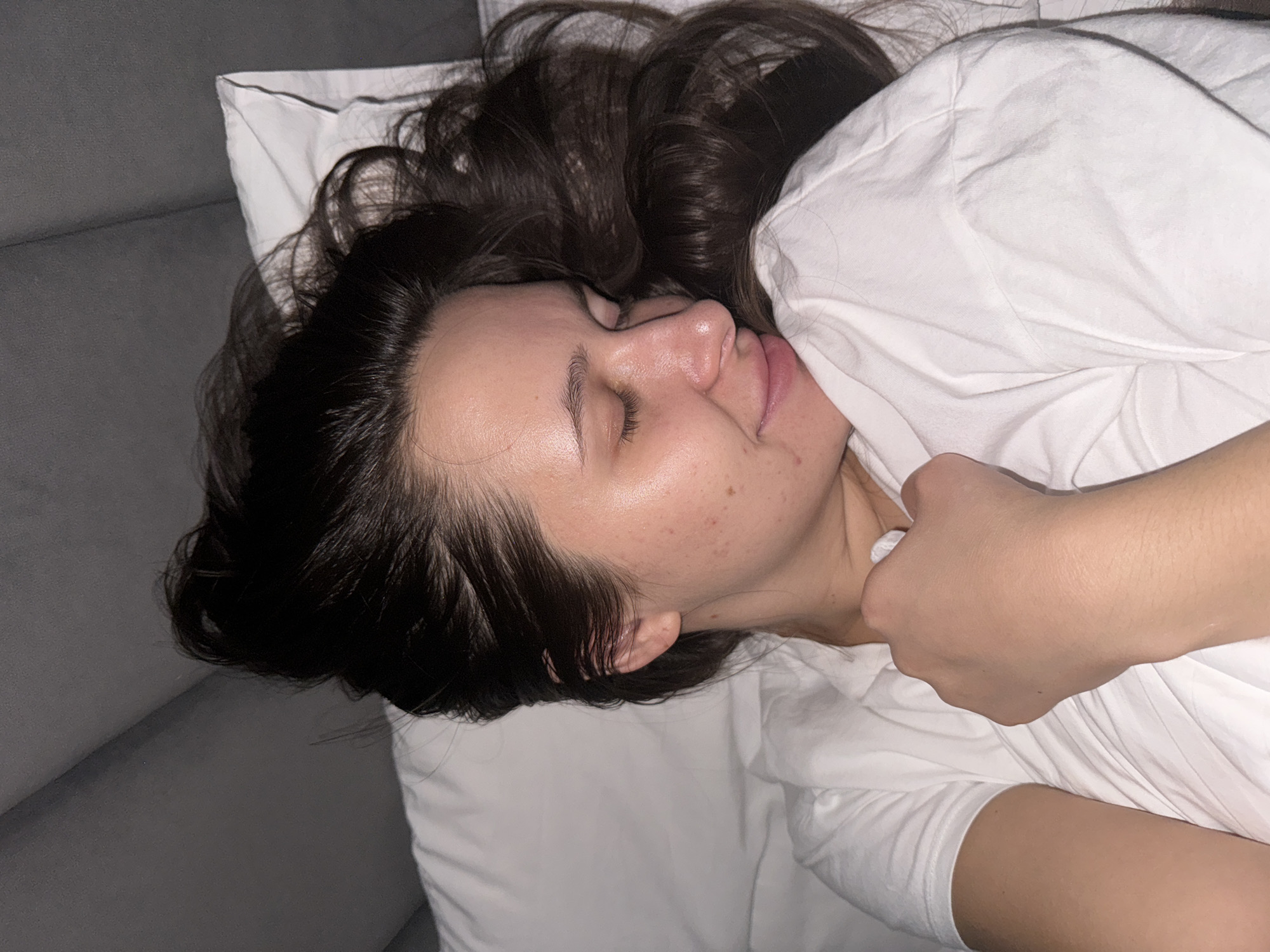 A woman with long, dark hair is lying on her side on a white pillow and white bedding. The background includes a gray surface, possibly a couch. The location is not discernible from the image. The image does not indicate any information specific to Akulovo or Kubinka, Russia.