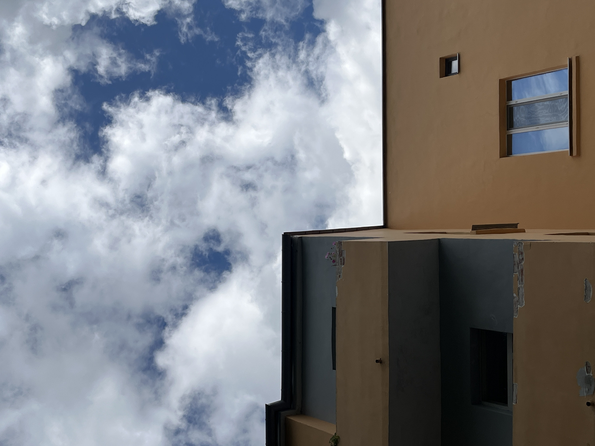 The image shows an upward view of two adjacent building facades against a sky filled with white cumulus clouds against a blue sky. The building on the left has a darker gray upper section with a protruding balcony. The balcony's underside is dark gray, and the front edge appears to be partially damaged, revealing brickwork. There are no people visible. The building on the right is a lighter tan color and features two windows. The larger window below is a multi-paned rectangular window reflecting the sky. A smaller, square window is located higher up on the right side of this facade. The architectural style and materials suggest an urban environment. This scene is not specifically identifiable as being in Vitsebsk, Belarus.