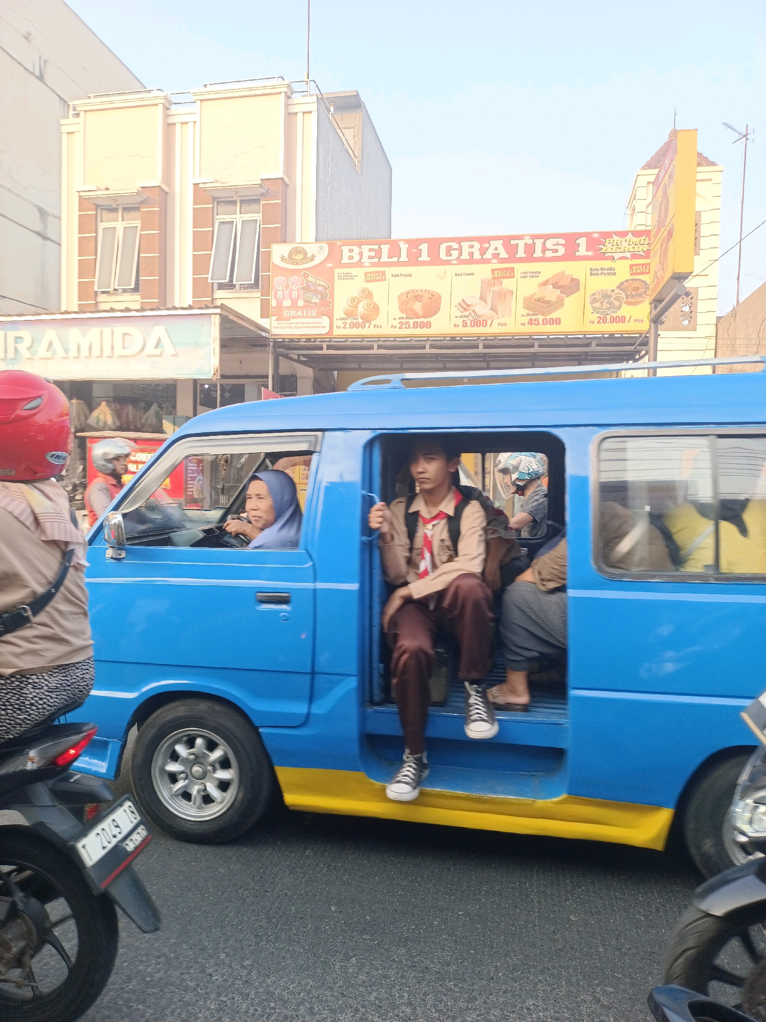 The image shows a blue minibus with its side door open, filled with passengers. A woman wearing a hijab is driving the minibus, and a young person in a scout uniform is seated in the open doorway. Other passengers are visible inside the minibus, some partially obscured. The minibus is driving on a paved road, with another motorcycle to the left of the frame. In the background, there are buildings, including a two-story building with arched windows and a billboard advertising a "Buy 1 Get 1 Free" promotion for various baked goods. The billboard is in Indonesian and displays prices in Indonesian Rupiah. The scene appears to be set in Karawang, Indonesia, a city known for its industrial areas and agricultural production, often with busy street life. The overall impression is a snapshot of daily life and transportation in a busy urban environment.
