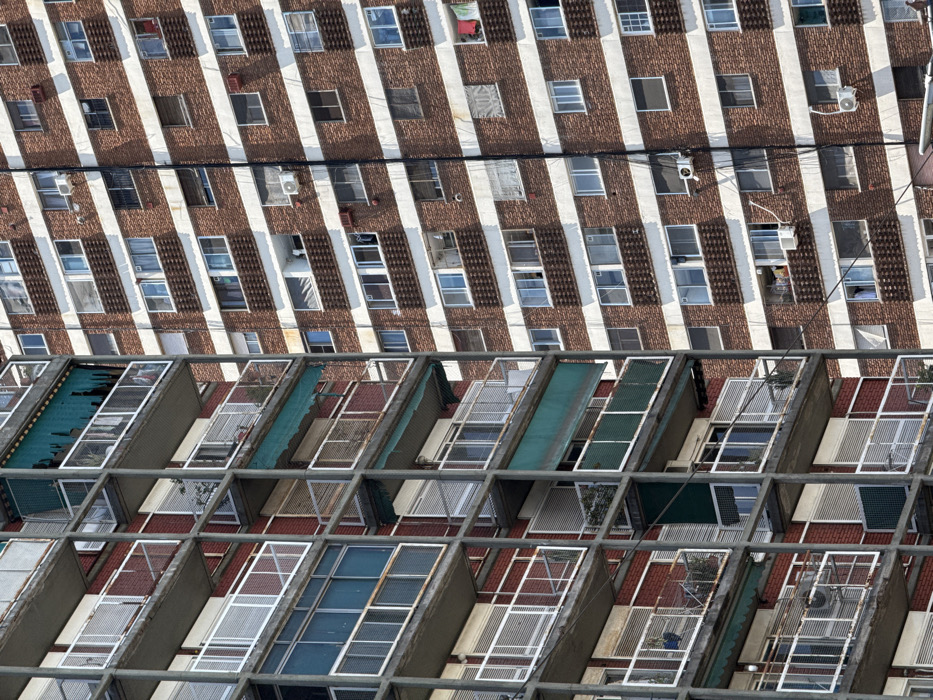 A high-angle shot reveals rows of balconies with diverse enclosures: mesh, slatted, and open designs. Some sport corrugated metal roofing. Above, a building facade features a repeating pattern of brick and vertical white stripes, punctuated by windows and air conditioning units. The perspective creates a strong diagonal emphasis within the frame.