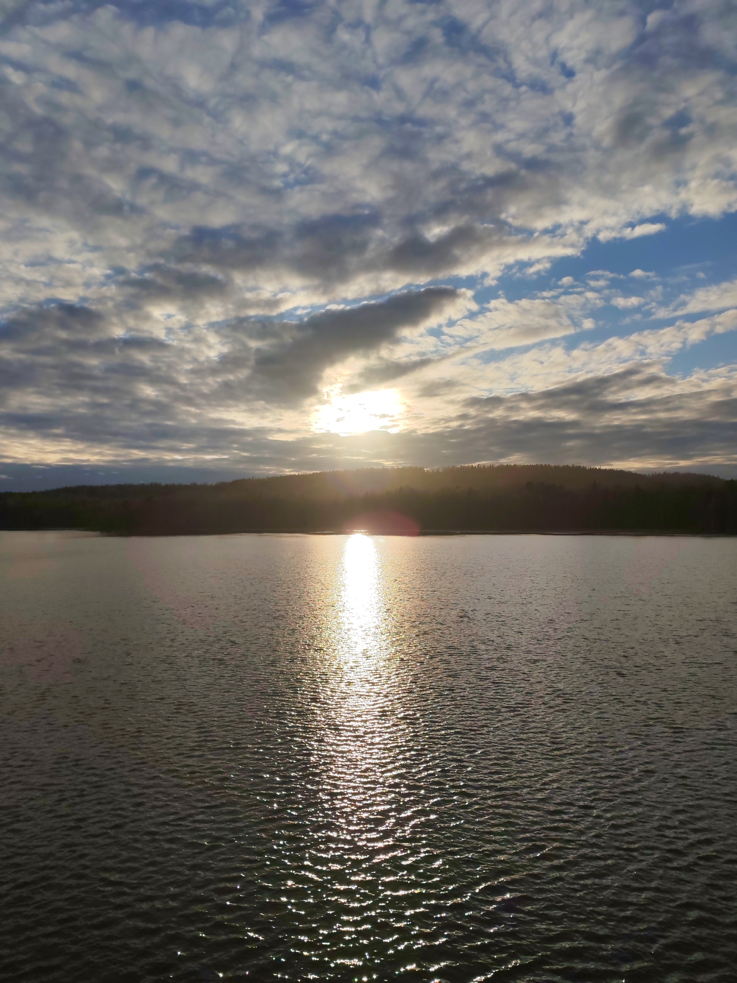 The image captures a serene landscape featuring a vast body of water under a partially cloudy sky, likely depicting a sunrise or sunset. The sun is low on the horizon, partially obscured by clouds, but its bright rays break through, creating a brilliant, shimmering path of light across the water towards the viewer.
The sky is filled with a mix of stratocumulus and altocumulus clouds, displaying various shades of grey, white, and hints of blue. Patches of clear blue sky are visible between the clouds, particularly towards the left and upper right of the frame. The overall lighting suggests either early morning or late afternoon, with a warm, golden glow emanating from the sun.
The body of water, which appears to be a large lake or reservoir, dominates the lower two-thirds of the frame. Its surface is rippled by gentle waves, enhancing the sparkling effect of the sun's reflection. The water gradually darkens towards the foreground where the sunlight doesn't directly hit.
In the mid-ground, a dark, tree-covered shoreline stretches horizontally across the image, forming a silhouette against the brighter sky and sun. The trees appear dense, suggesting a forested area. No people or man-made structures are visible.
The scene conveys a sense of natural beauty and tranquility, characteristic of a remote landscape like Shcheleiki, Russia. There is no visible text in the image.