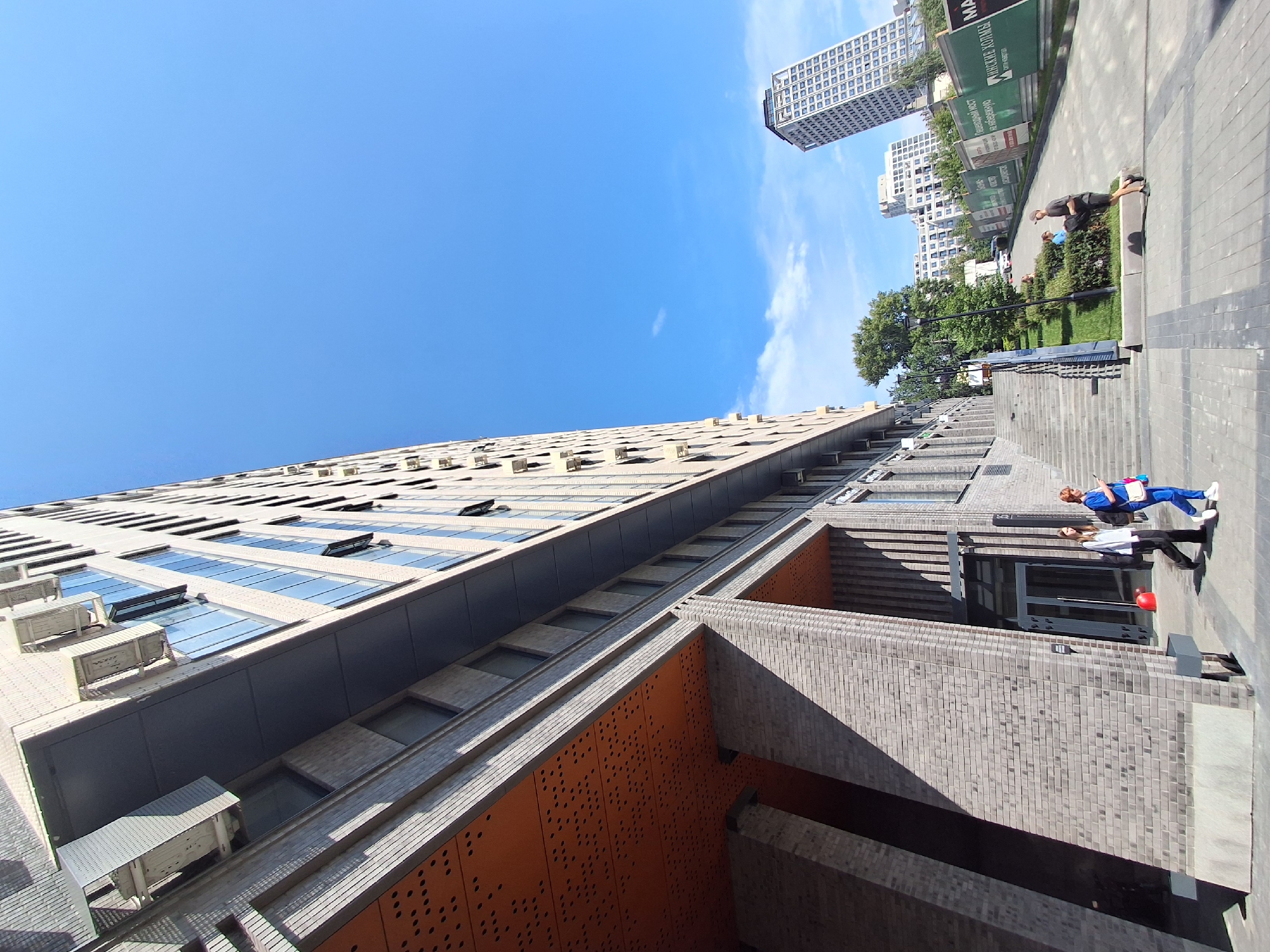 The image shows a modern building with brick and glass facades, likely an apartment or office complex, in Novosibirsk, Russia. The building is tall and angular, with several balconies and air conditioning units visible. The sky is clear blue with a few scattered clouds. In the foreground, there are people walking on a paved sidewalk. One person is wearing a blue uniform and carrying a bag, while another is in casual clothing. There are also trees and greenery in the background, suggesting an urban park or landscaped area. Further in the distance, other buildings and a tall skyscraper can be seen. The overall scene depicts a bright, sunny day in a contemporary urban environment.