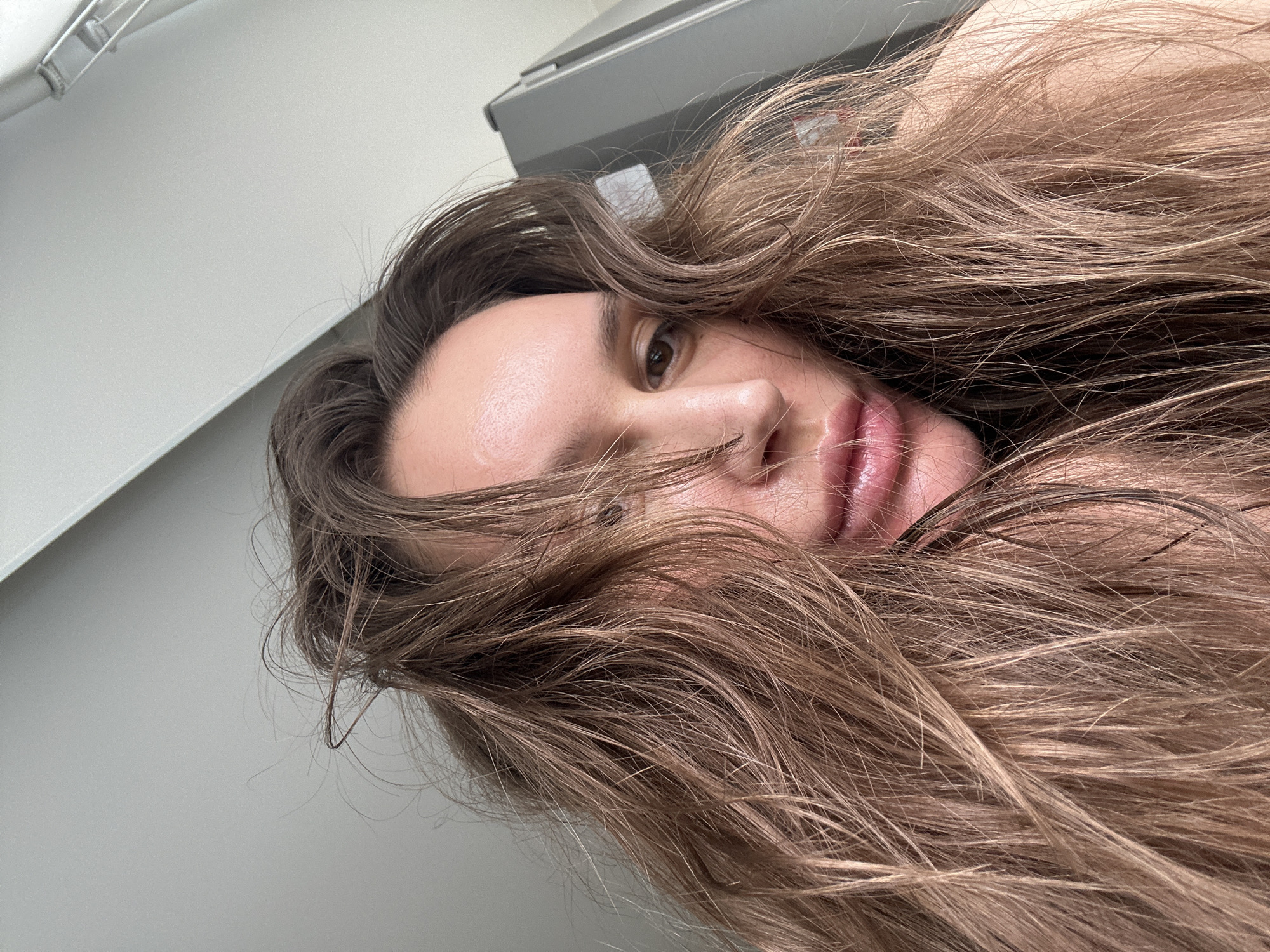 The image is a close-up selfie of a person with long, wavy brown hair partially covering their face. The person's eye, nose, and lips are visible. The background is a plain white or light grey surface. There is no information in the image to indicate a location in Moscow, Russia, or any other specific city.