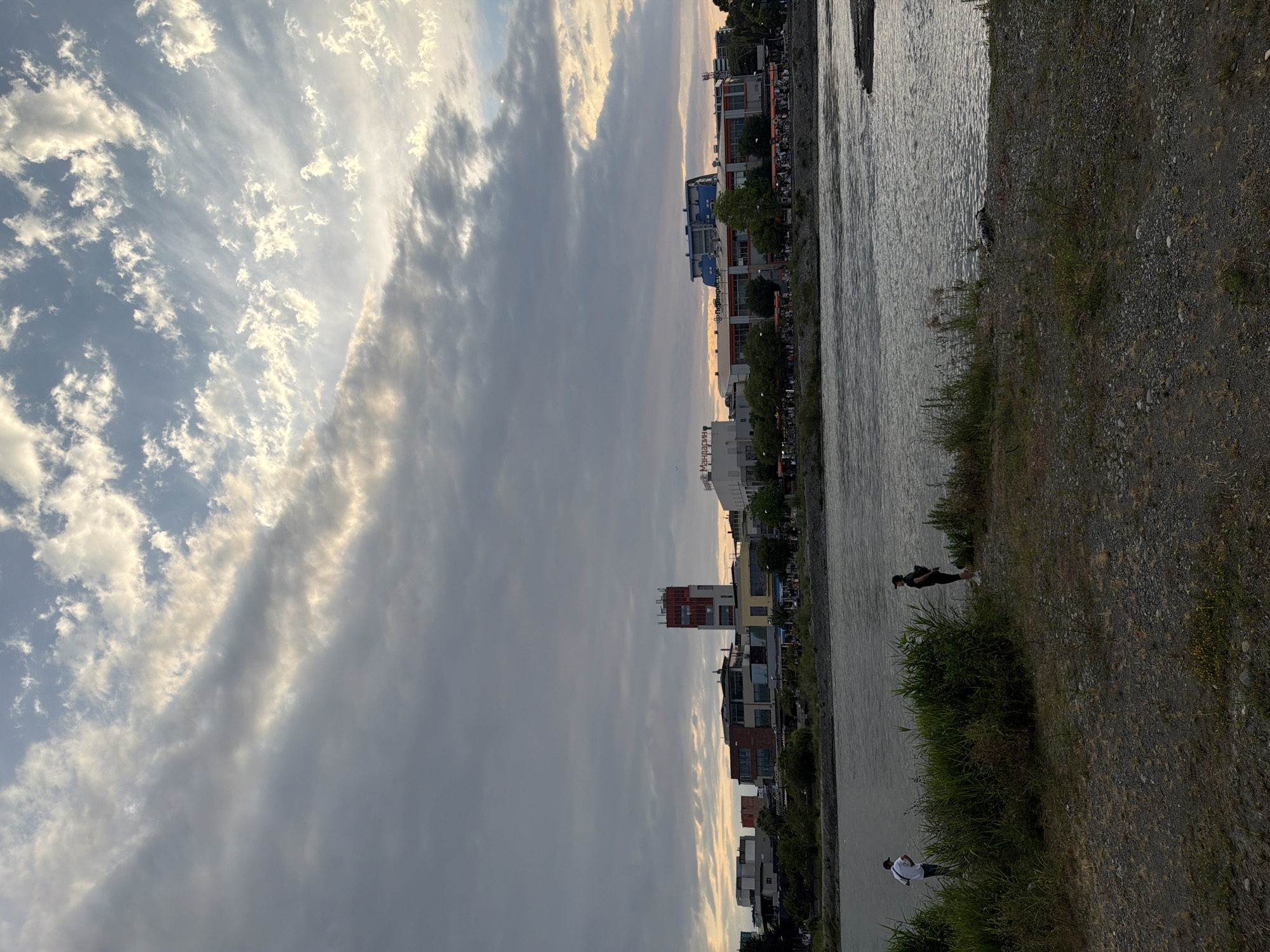 This image depicts a view of the city of Sochi, Russia, across a wide river. The sky is filled with dramatic clouds, some illuminated by the setting sun, creating a mix of dark and light tones. In the foreground, there is a gravelly shore with some vegetation. Two individuals are visible on the left side of the riverbank, one standing and facing away from the camera, and the other facing towards the river. Across the river, a cityscape unfolds with various buildings, including a prominent red and white tower. Shops and other commercial establishments line the waterfront, with trees and people visible along the promenade. The river itself appears calm, with ripples reflecting the sky. The overall scene suggests a late afternoon or early evening setting.