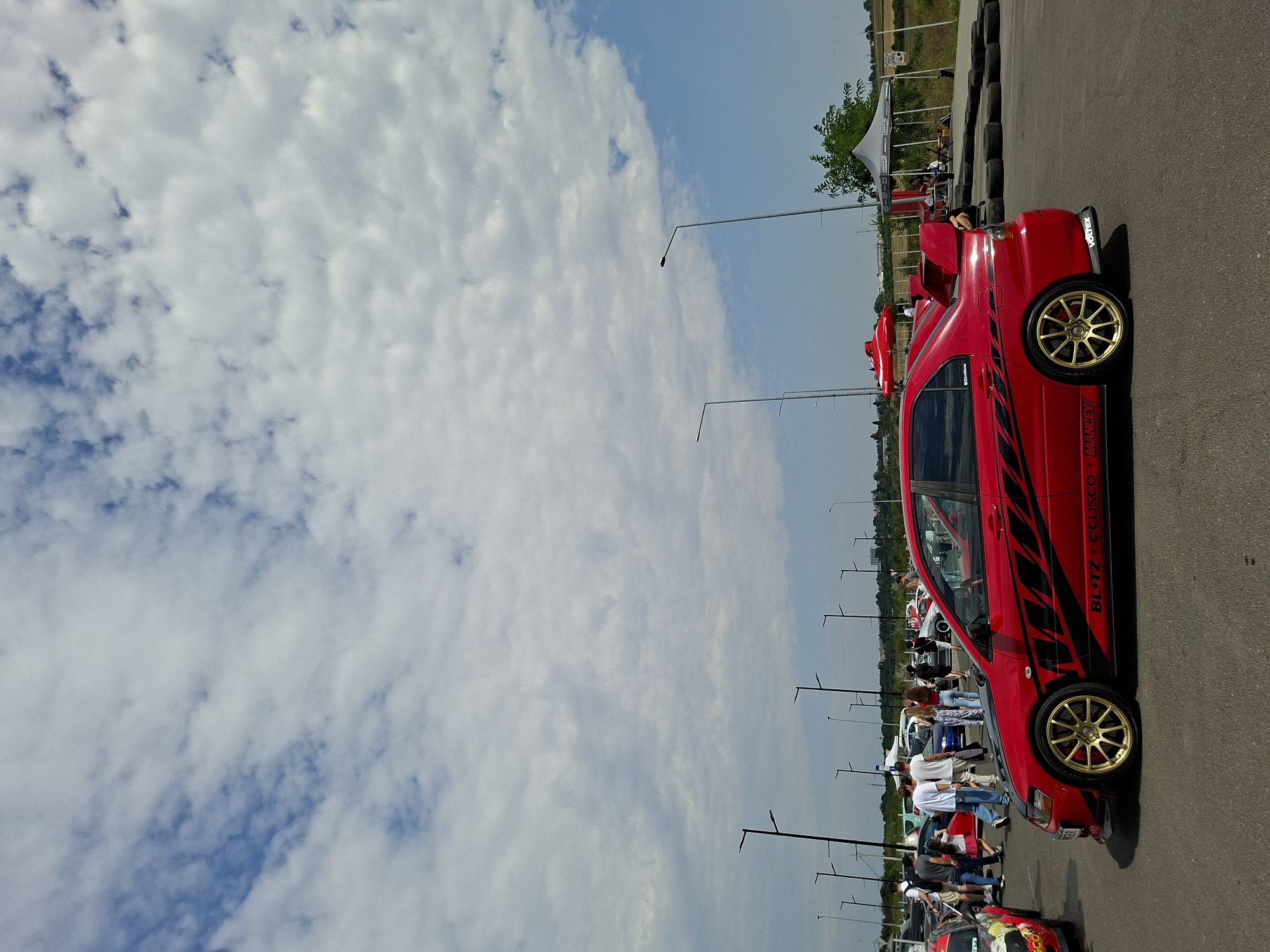 The image depicts an outdoor event, likely a car show or race gathering, at the Stăuceni Aerodrome in Stăuceni, Moldova. The foreground is dominated by a bright red Mitsubishi Lancer Evolution, characterized by its aggressive body kit, large rear spoiler, and gold rims. The car features black racing decals and visible branding such as "BLITZ" and "CUSCO". To the left of the red car, another vehicle with colorful racing livery is partially visible.
A crowd of people, casually dressed in summer attire, is gathered behind the vehicles, suggesting active participation or spectating. Several tall lampposts are strategically placed throughout the paved area. In the background, tents are set up, one of which appears to be a merchandise or information booth. Beyond the event area, a rural landscape with fields and trees is visible under a partly cloudy sky. The overall scene conveys a sense of organized activity and a warm, bright day.