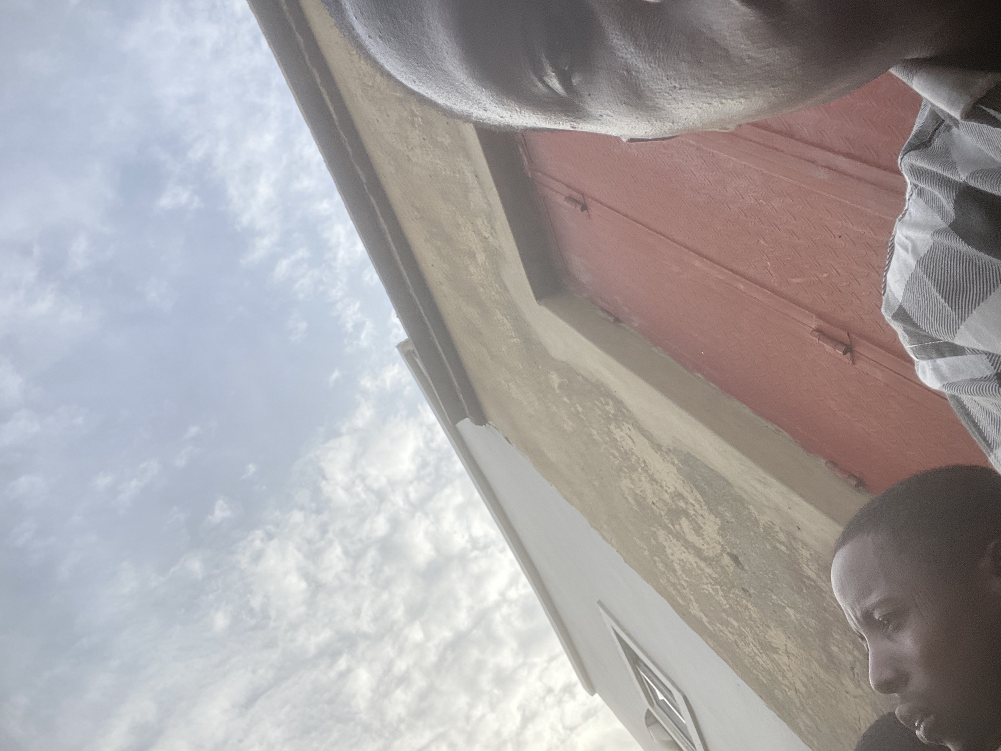 A low-angle shot depicts two individuals, presumably African-American males. The head of one is prominently featured in the upper portion of the frame, seemingly taking the photograph. A portion of the other person's head and shoulders are visible in the lower right corner. The background includes part of a building with a red door and a cloudy sky. The location is Unknown, Unknown.