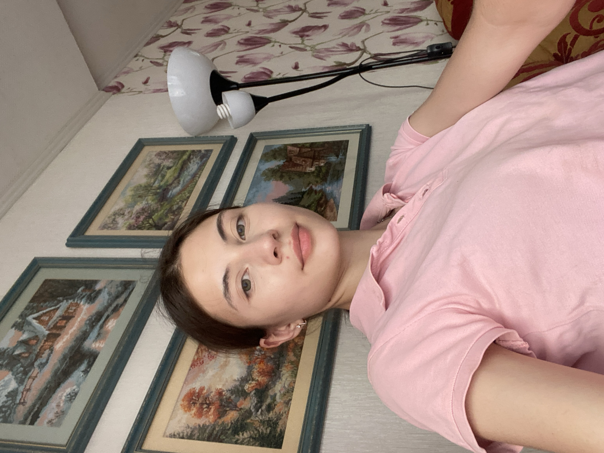 A young woman with brown hair and fair skin is shown in a close-up selfie. She is wearing a pink polo shirt and has a small hoop earring in her left ear. The background features three framed landscape paintings on the wall and a floor lamp with a white shade. To the right, floral wallpaper is visible. The setting appears to be a residential interior in Votkinsk, Russia.