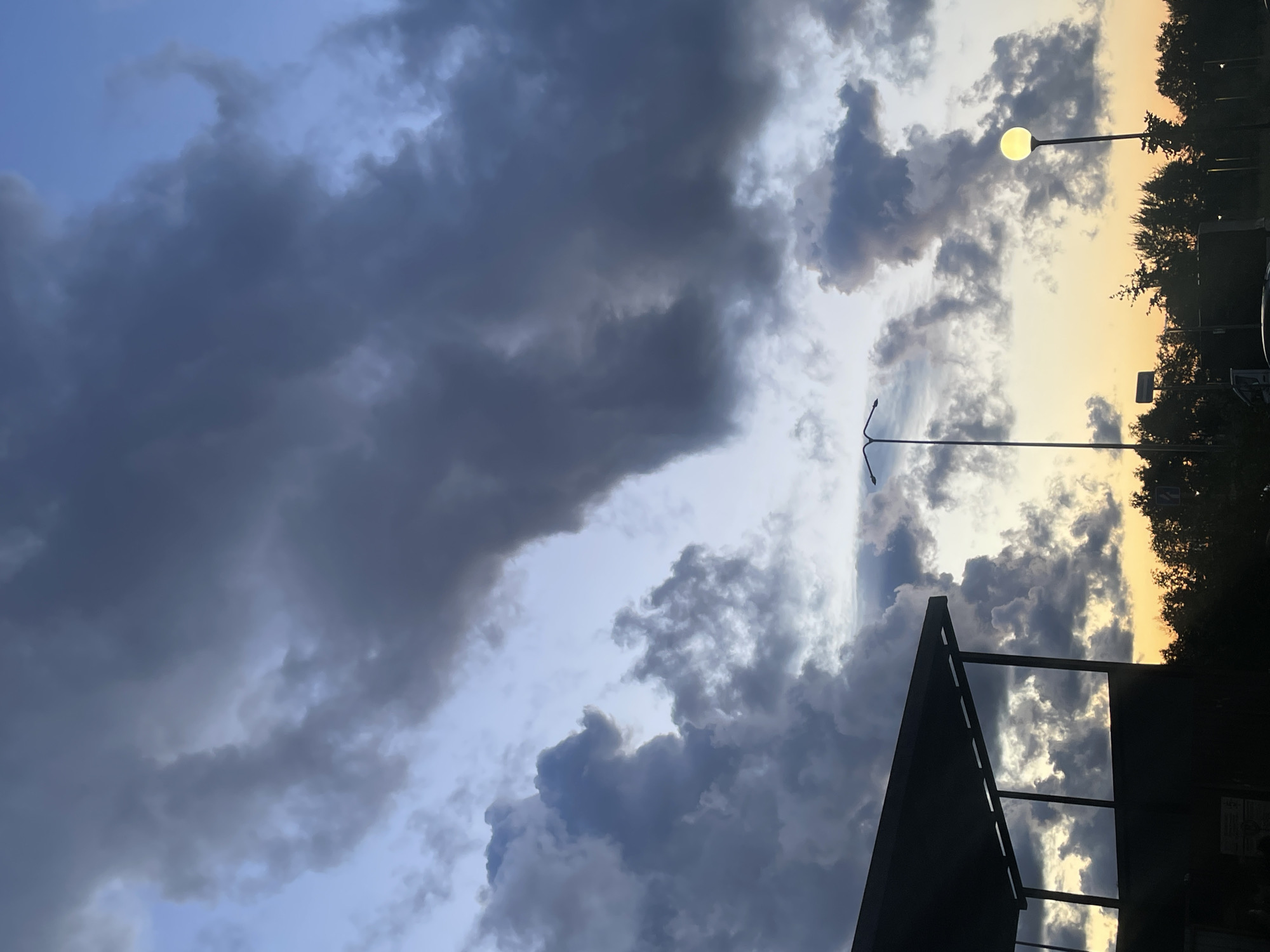 The image depicts a dramatic sky filled with dark, heavy clouds illuminated by the fading light of sunset. In the foreground, a silhouetted structure, possibly a bus stop or a shelter, occupies the left side of the frame. A street lamp with a bright, round bulb stands to the right, while another, more modern street light with a longer arm is visible behind it. The horizon is lined with dark silhouettes of trees, suggesting an urban or suburban edge. The sky transitions from a pale blue at the top to a warm yellow and orange hue near the horizon, where the sun is setting. The overall mood is atmospheric and slightly ominous due to the presence of the storm-like clouds. The location is identified as Saint Petersburg, Russia.