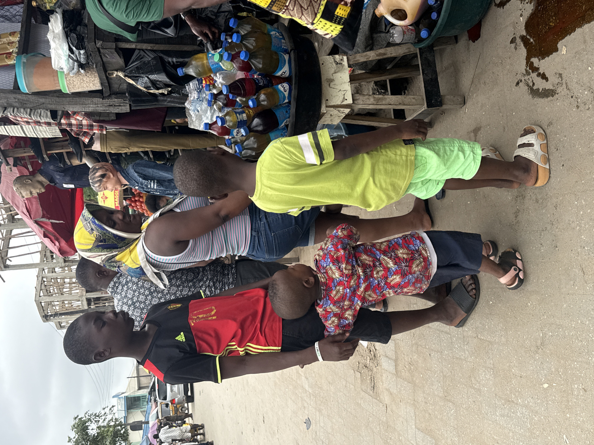 The image depicts a street scene in Ado, Nigeria, featuring a group of people near a market stall. In the foreground, a young boy wearing a bright green t-shirt and light green shorts stands with his back to the camera, looking towards a stall laden with bottles. To his left, an older boy in a red and black football jersey and black shorts stands, holding the younger boy's hand. A woman in a striped tank top and blue denim shorts, wearing a headscarf, is also visible. The market stall displays various bottled drinks and other goods. In the background, other individuals and market activities are suggested. The environment appears to be an outdoor marketplace with a rough, paved ground.