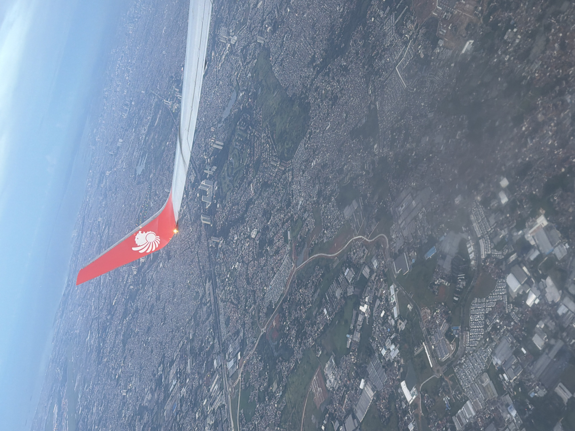 The image shows an aerial view from an airplane over Ciakar, Indonesia. Visible is the wing of the plane, bearing a red color and the logo of a lion. Below, the landscape is dominated by a dense urban area, characterized by buildings and infrastructure. The horizon line is visible in the upper left. The overall impression is one of a densely populated area.