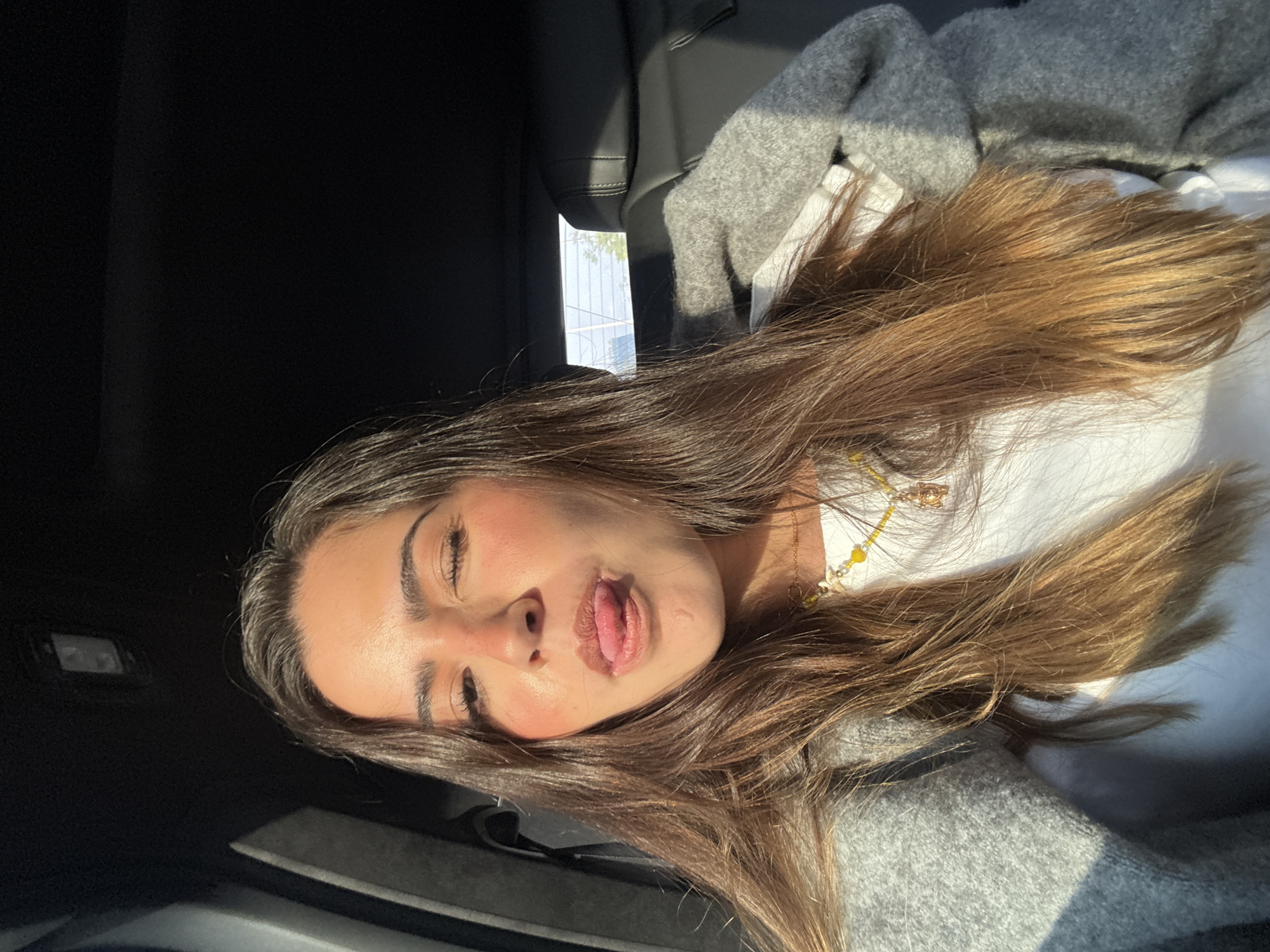 A young woman with long brown hair is in the back seat of a car, sticking her tongue out and squinting her eyes. She is wearing a white shirt, a gray sweater, and a necklace with yellow beads. The location is San Bernardino Tlaxcalancingo, Mexico. Sunlight is shining through the car window.