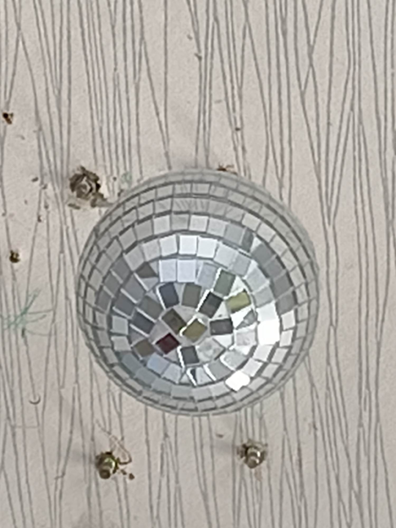 A disco ball hangs from a light-colored wall with a vertical stripe pattern. The disco ball is covered in small, square mirrored tiles, reflecting light. There are several small pieces of debris on the wall, including some dark, textured clumps near the top left and some lighter, smaller specks scattered around. Below the disco ball, two small, metallic objects are attached to the wall, resembling screws or anchors. The overall scene is static and appears to be an indoor setting, likely a domestic or event space. There is no indication of its specific location in Moscow, Russia, or any people present.