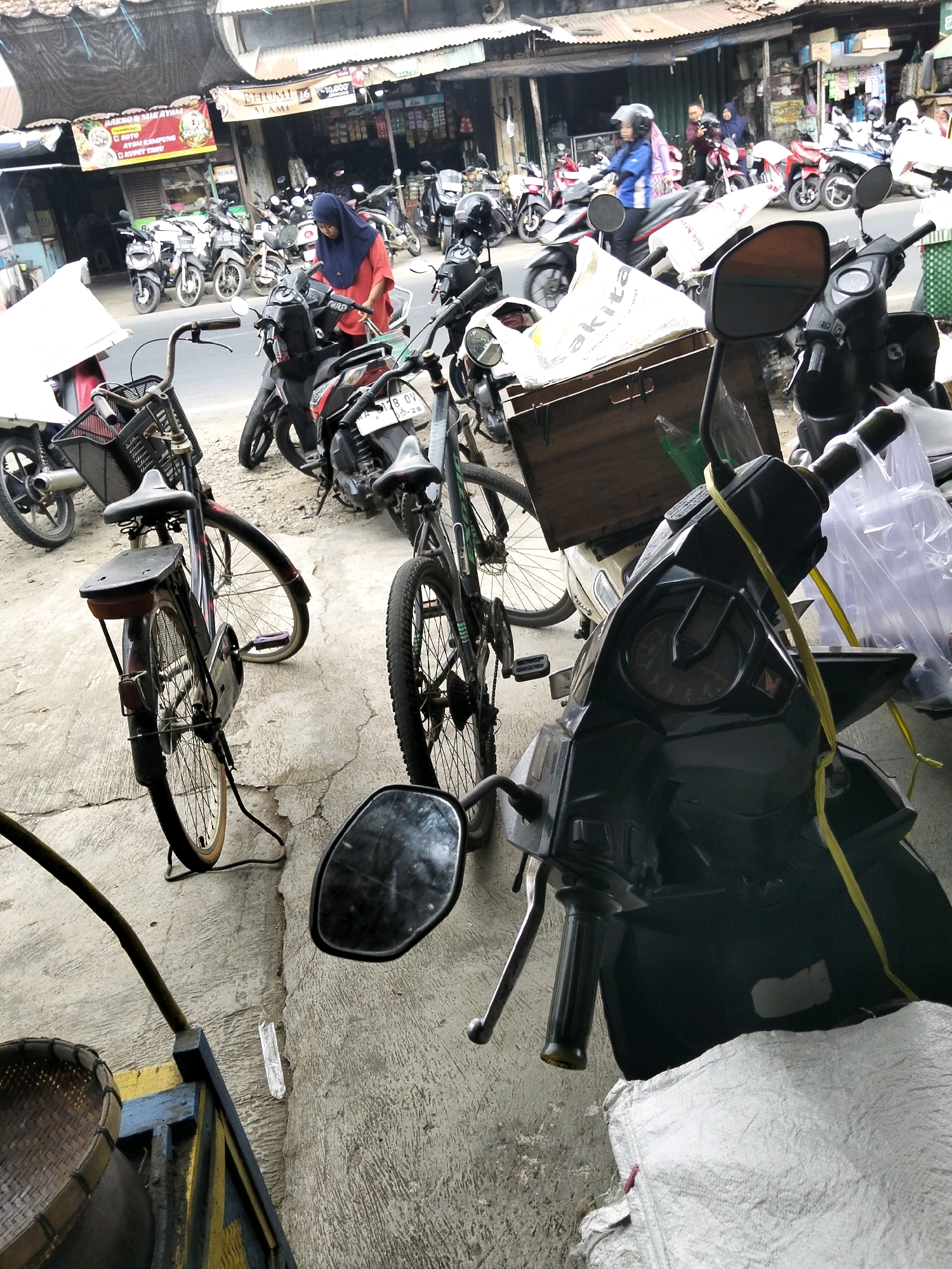 The image depicts a street scene in Kemiri Kidul, Indonesia, with a focus on parked motorcycles and bicycles. In the foreground, a black scooter is prominent, its handlebars and front panel visible. A yellow tie is looped around the handlebars. To the left, a vintage bicycle with a basket and a thin frame is parked, its rear wheel slightly splayed. Behind it, another bicycle, also with a basket, leans against a wooden crate. Further back, a woman in a red shirt and hijab is standing next to a motorcycle, appearing to interact with it. Several other motorcycles are parked in rows, some with helmets on their seats or handlebars. The background shows storefronts with awnings and various goods displayed, typical of a market or street vendor area. The ground is paved with concrete and some dirt. The overall atmosphere suggests a busy commercial area with a strong presence of two-wheeled transportation.