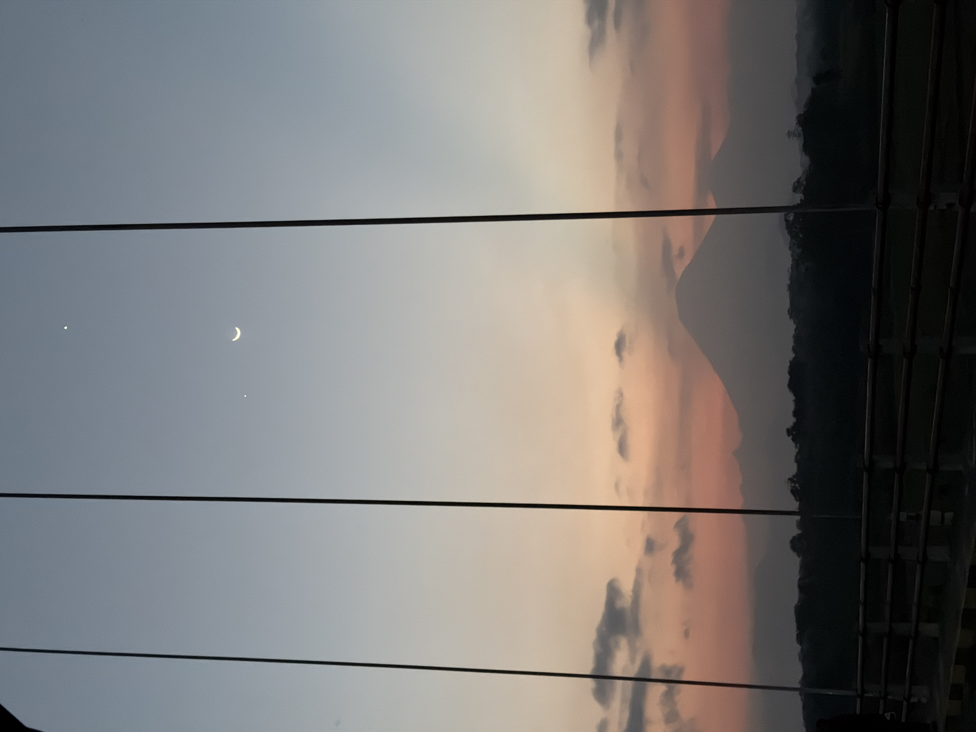 The image depicts a sunset view from a bridge in Selokanayar, Indonesia. The sky transitions from a pale blue at the top to shades of pink and orange near the horizon, with scattered clouds. In the upper sky, a crescent moon and a small bright star are visible. Silhouetted against the colorful sky are two prominent mountains in the distance, appearing as dark, conical shapes. Below the mountains, a dense line of trees forms the horizon. The foreground of the image shows the structure of the bridge, with dark vertical cables and a railing. The railing is made of dark horizontal bars and has concrete supports. The overall scene is serene and atmospheric, characteristic of a twilight view.