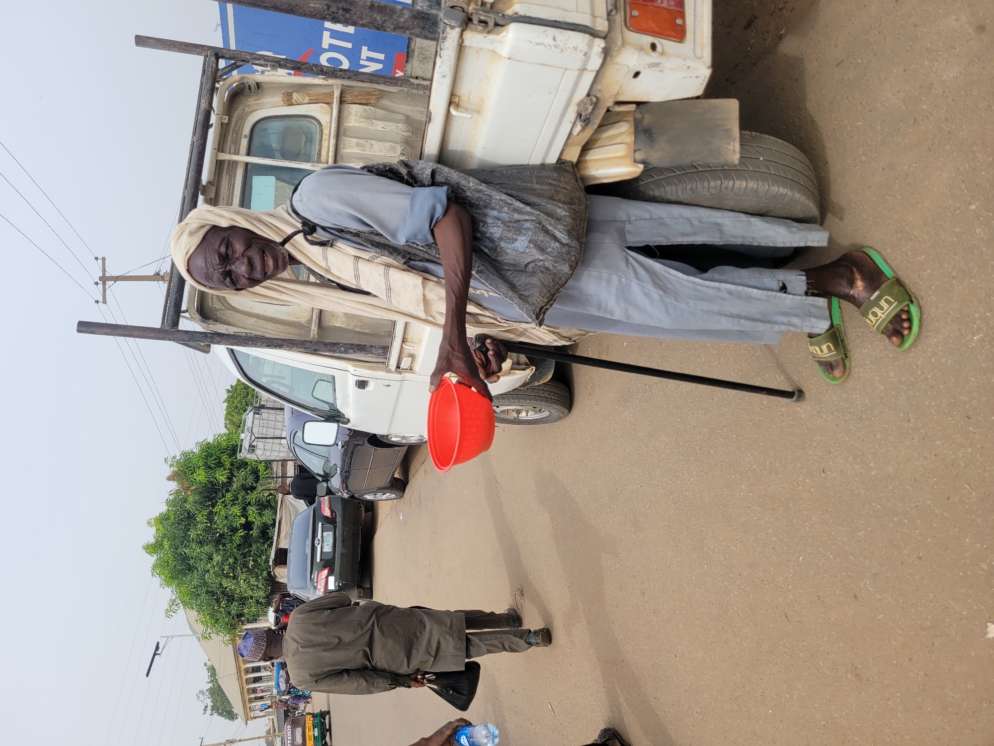 This media file captures an outdoor street scene in what appears to be an urban or semi-urban environment in Jos, Nigeria, during the daytime with an overcast sky.
The central figure is an elderly man with dark skin, positioned on the right side of the frame. He wears a light beige head covering, a light blue long-sleeved shirt, light grey trousers, and green slide sandals branded with "unbun." A dark, patterned garment is draped over his shoulders. He holds a red plastic bowl in his right hand and a black walking stick in his left. He has a pleasant expression, with a noticeable smile. He is standing next to a white pickup truck, which has a partial blue sign on its side showing the text "OTE NT".
In the foreground, another person is seen from behind, wearing a dark coat and trousers, walking away from the camera. A hand in the bottom right corner holds a plastic water bottle. The ground is a paved, light brown surface.
The background features several vehicles, including a black sedan, parked or moving along the street. Lush green trees and some distant buildings are visible. Overhead, power poles and lines stretch across the frame against the bright, hazy sky. The overall scene depicts a common street vista, possibly near a market or a busy thoroughfare.