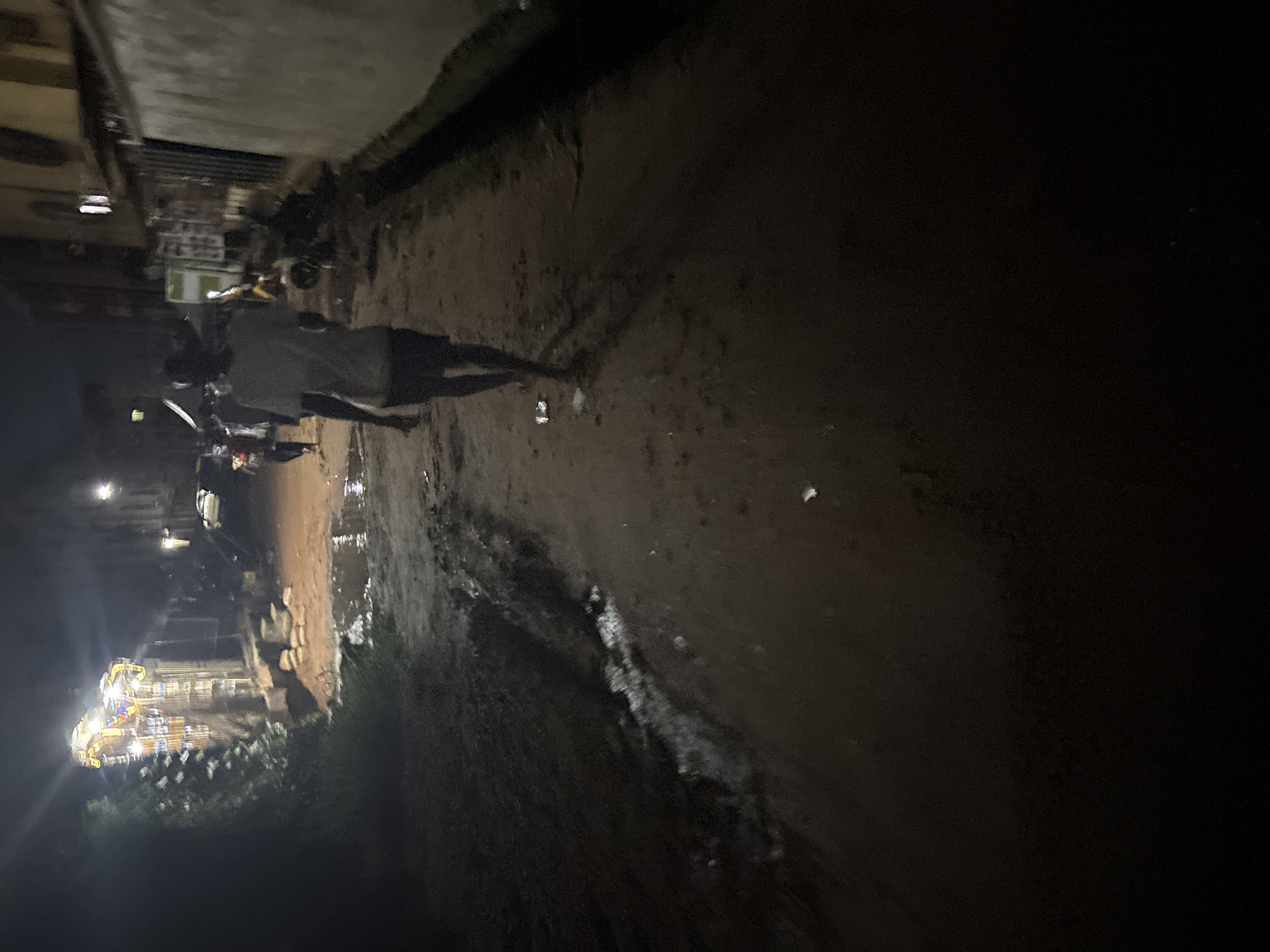 The image depicts a dimly lit street scene in Onitsha, Nigeria, at night. A lone individual, likely a man, walks away from the camera down a dirt path that is uneven and shows signs of recent rainfall with puddles of water present. The person is wearing a light-colored t-shirt and dark shorts. In the background, there are buildings and some festive lights are visible on structures to the left, suggesting a possible event or market area. The overall environment appears to be a less developed urban area, with a mixture of natural elements and man-made structures. The lighting is poor, casting long shadows and obscuring some details of the surroundings.