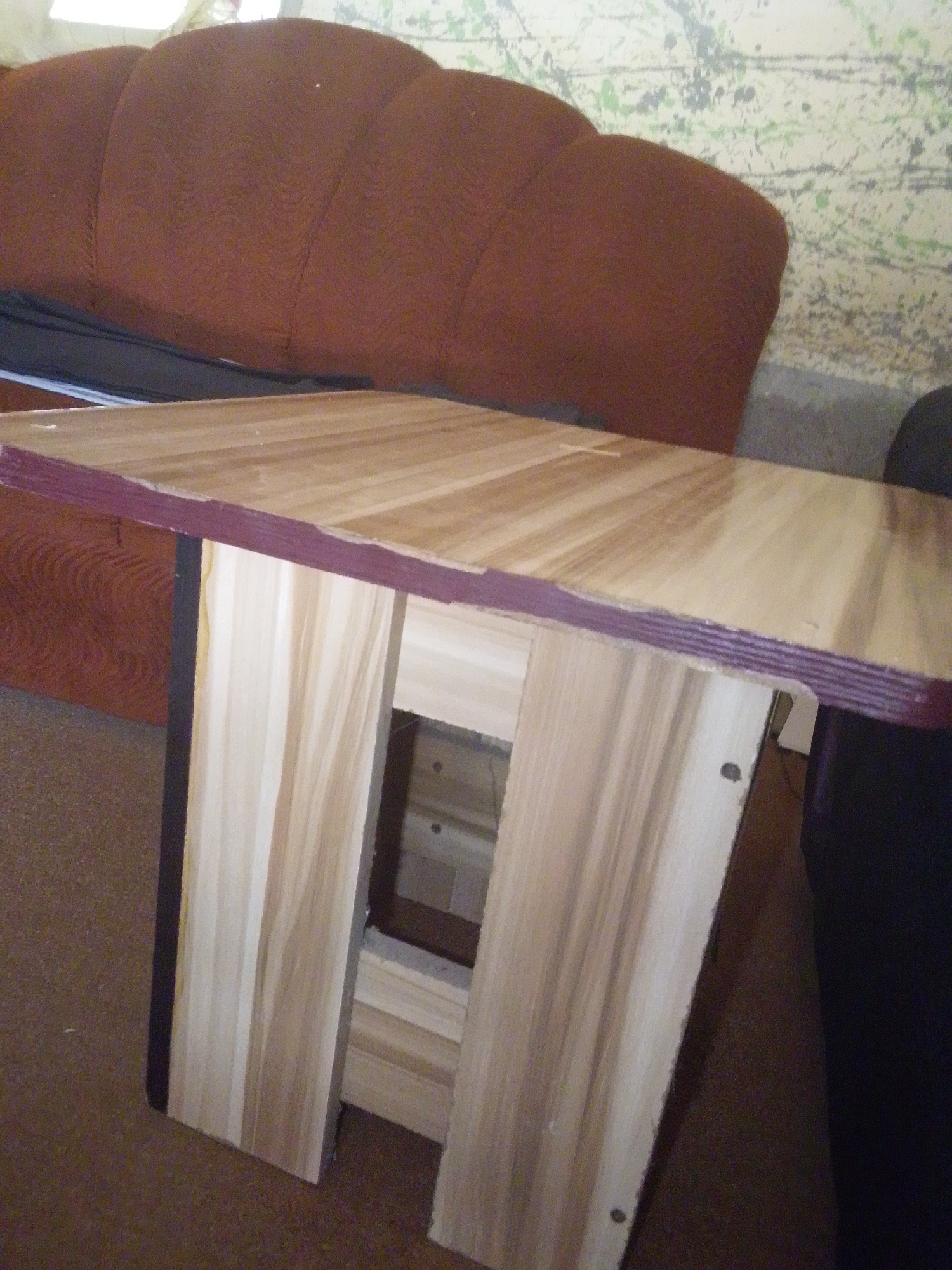 The image shows a close-up of a handmade wooden table with a light wood grain, possibly pine or a similar light-colored wood. The tabletop has a dark reddish-brown edge. The table has two vertical supports made of the same light wood, connected by horizontal pieces to form an open rectangular space in the center. The table is placed in what appears to be an indoor setting. In the background, a large, plush, dark brown sofa is visible, with a wall behind it decorated with a mottled pattern of green, gray, and black splatters, resembling abstract art or a distressed wallpaper. A dark object, possibly a chair or another piece of furniture, is partially visible on the right side of the image. The lighting suggests it is daytime, but the exact time is not discernible. There are no people visible in the image. The overall scene suggests a domestic or casual environment. The location context of Jalingo, Nigeria, provides a geographical anchor but does not offer specific visual cues within the image itself to confirm this location. No visible text is present.