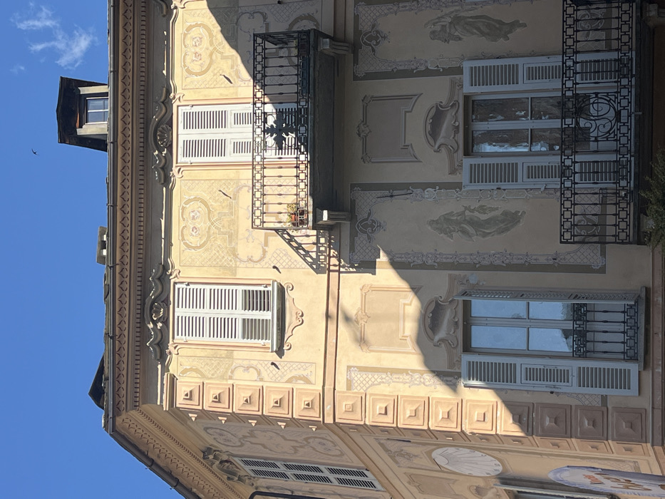 A multi-story building facade in Varallo, Italy, is presented under clear blue skies with strong, directional sunlight. The structure exhibits a light peach-colored exterior, extensively decorated with painted frescoes featuring geometric patterns, ornate scrolls, and large-scale figural depictions, including a classical female figure on the lower level. The building's corner is articulated with prominent, raised square quoins. Multiple rectangular windows, each equipped with white louvered shutters, are visible; some shutters are closed, while others on the lower floor are open, revealing reflective glass panes. Two black wrought-iron balconies extend from the facade; the upper balcony holds a small potted plant and casts a distinct, elaborate shadow onto the wall below. A partial sundial with a shell design is visible on the far-left corner. A small, dark bird is silhouetted against the blue sky in the upper central portion of the image. Sharp shadows from architectural features and an off-camera element, possibly foliage, are cast across the sunlit facade.
