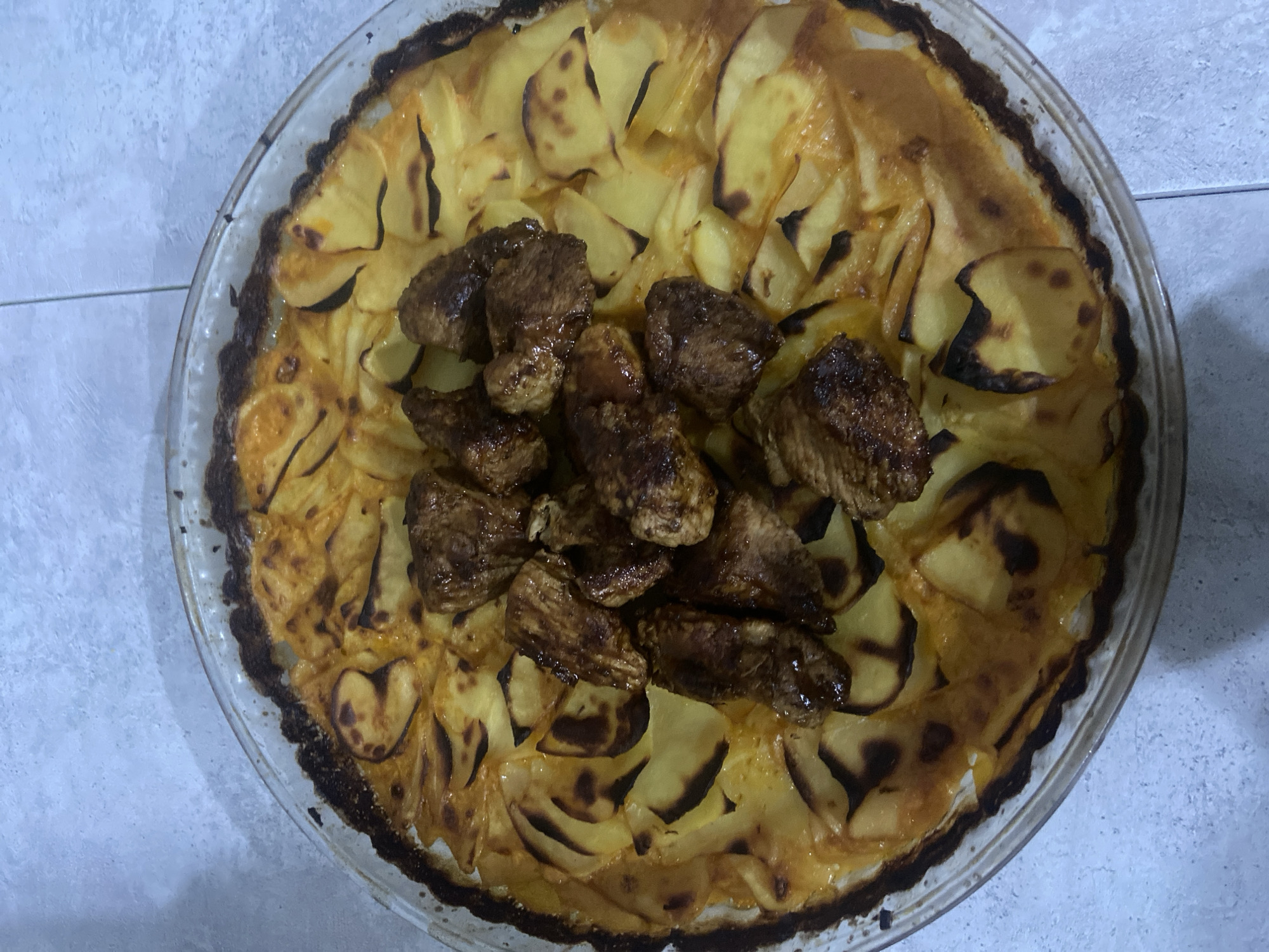 A glass baking dish containing a baked dish is positioned centrally on a light grey tiled surface, likely a floor or countertop, in Chaikovsky, Russia. The dish consists of a base of thinly sliced, overlapping potatoes that are golden-brown and partially charred at the edges, arranged in a circular pattern. A cluster of approximately ten to twelve irregularly shaped pieces of cooked meat, appearing dark brown and somewhat glossy, are placed in the center of the potato base. The glass dish has a scalloped edge and appears slightly burnt along the rim where it meets the potato layer. The overall lighting is even, with a slight overhead light source. No people or actions are visible.