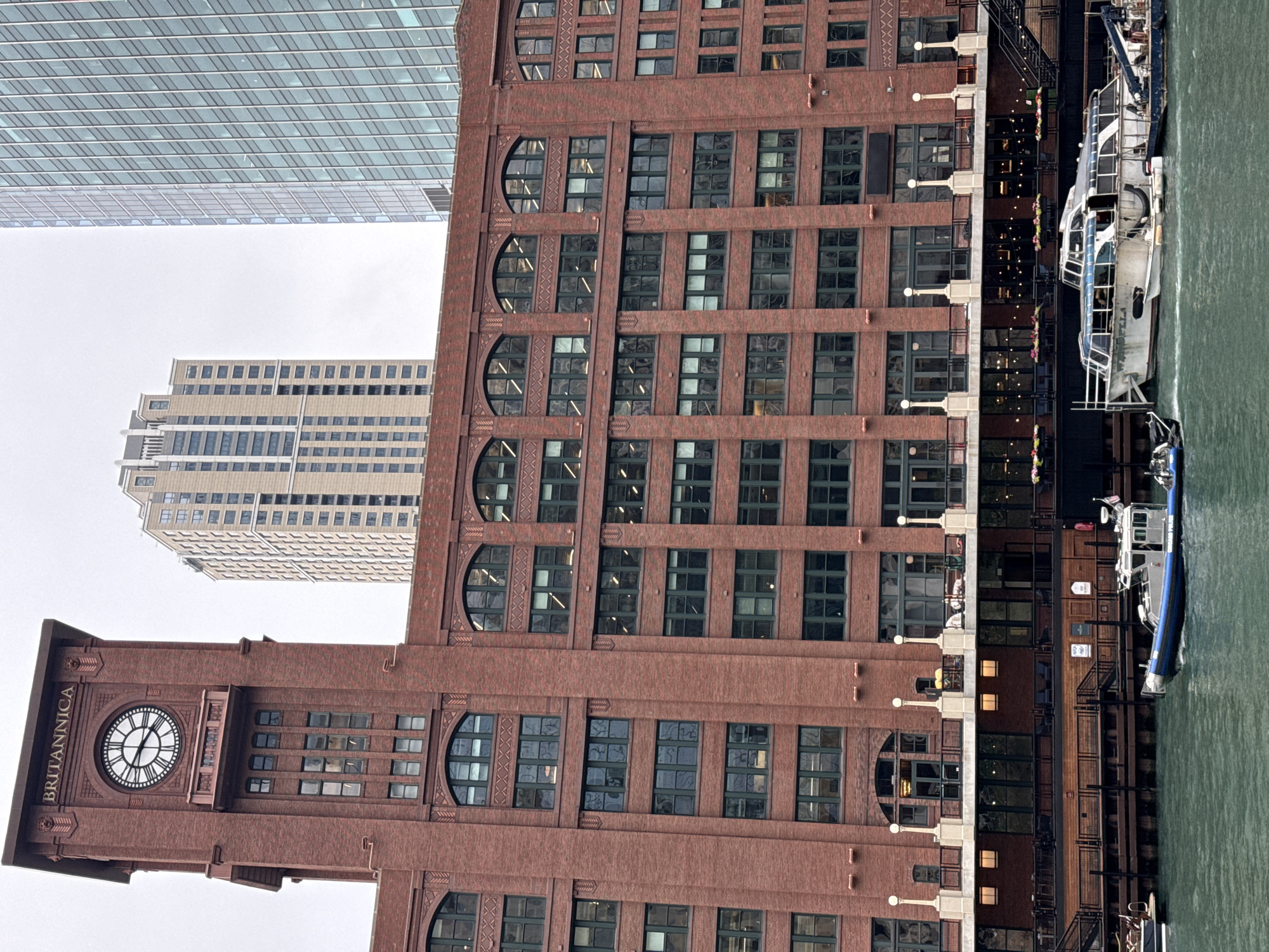 This is a photograph taken in Chicago, United States of America, featuring a cluster of buildings along a waterfront. The dominant structure is a red brick building with a clock tower. The clock face on the tower is clearly visible and bears the inscription "BRITANNICA." The building has numerous windows, many of which reflect the overcast sky.
Adjacent to the red brick building is a modern, glass-covered skyscraper, its reflective surface creating a stark contrast to the historic architecture. Further in the background, another tall, lighter-colored building stands, also characterized by rows of windows.
In the foreground, the murky green water of a river or canal is visible, with a white boat docked alongside the red brick building. The boat appears to be a tour boat or ferry, with multiple decks and windows. Some people are visible on the lower deck of the building, suggesting it might be a restaurant or public space.
The sky is overcast, indicating a cloudy or possibly rainy day. The lighting suggests it is daytime. The overall scene conveys an urban setting, likely a business or tourist district where historical and modern architecture coexist, with a waterway playing a central role in the landscape.