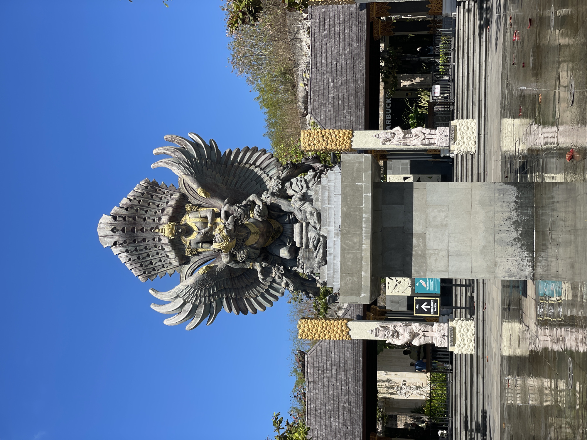 The image captures a wide, frontal view of the Garuda Wisnu Kencana (GWK) Cultural Park in Kuta Selatan, Indonesia. The centerpiece of the scene is a colossal statue of Garuda, a mythical bird-like creature from Hindu mythology, majestically perched on a pedestal. The statue is intricately detailed, with large wings spread wide, a bejeweled headdress, and a golden-colored adornment around its torso. Garuda is depicted in a seated position, holding onto a smaller figure below.
In front of the statue, a reflective water feature creates shimmering patterns on the paved ground, with a few fallen leaves scattered across its surface. The water shows reflections of the statue and the surrounding architecture. Behind the statue, a tiered structure with a dark, tiled roof extends outwards, flanked by two ornate pillars topped with golden, textured decorations.
To the sides of the main pathway, there are two stone sculptures of human figures, one on each side. The left sculpture is labeled "RAHWANA" and the right one "RAMA," both standing on decorative bases. Further back, a set of stone stairs leads up to the entrance of a building that appears to be a Starbucks, with its distinctive logo visible. People are seen walking on the stairs and near the entrance. The background consists of a sloping, rocky hill covered with vegetation. The sky above is a clear, vibrant blue. The overall scene is bathed in bright sunlight, casting distinct shadows on the ground.