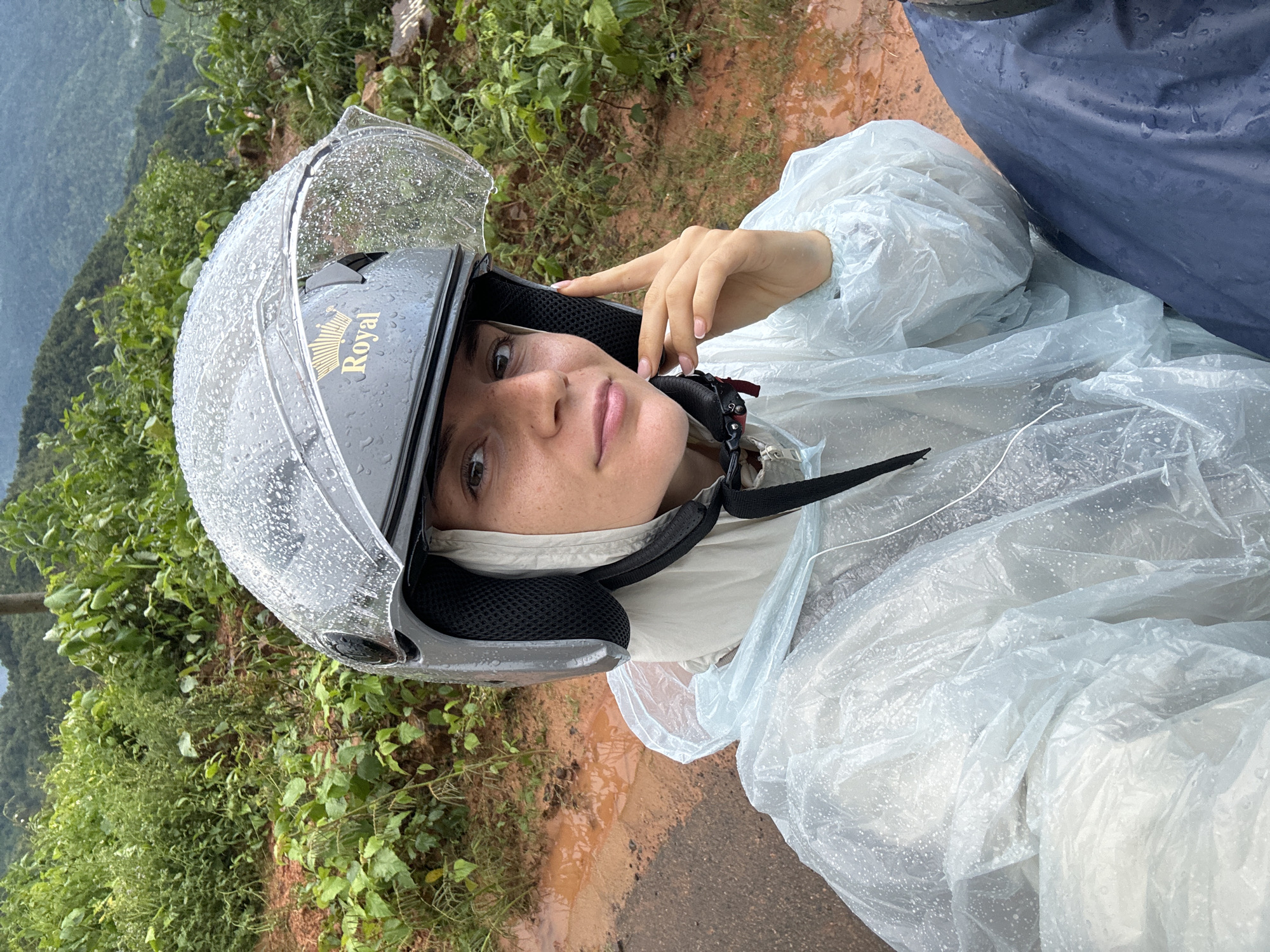 A close-up, over-the-shoulder selfie shows a woman wearing a helmet and a transparent raincoat on a motorcycle. The helmet is gray with a clear visor covered in raindrops. The word "Royal" is visible in gold on the side of the helmet. She is wearing a light-colored inner layer under the helmet and a light blue, slightly crinkled transparent raincoat. She is holding her right hand up to her chin, with her fingers lightly touching her face. The background consists of lush green vegetation and a muddy, wet ground, suggesting a rainy outdoor environment. The scene appears to be on a winding road in a hilly or mountainous area, characteristic of the landscapes found in Nao Sẻ, Vietnam.
