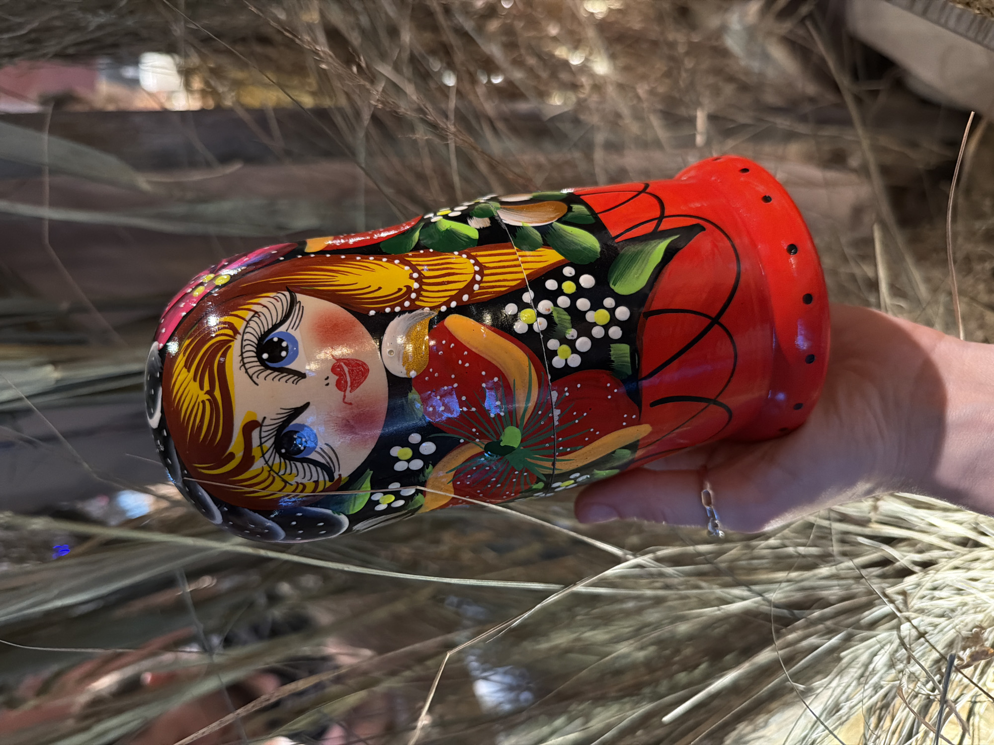 A close-up shot shows a hand holding a partially opened, brightly painted matryoshka doll against a blurry background of dried grass or hay. The doll is decorated with a traditional Russian floral design and the face of a woman with large blue eyes. The hand, wearing a delicate silver ring, suggests the doll is being displayed or offered. There is no indication in the image that it was taken in Khimki, Russia.