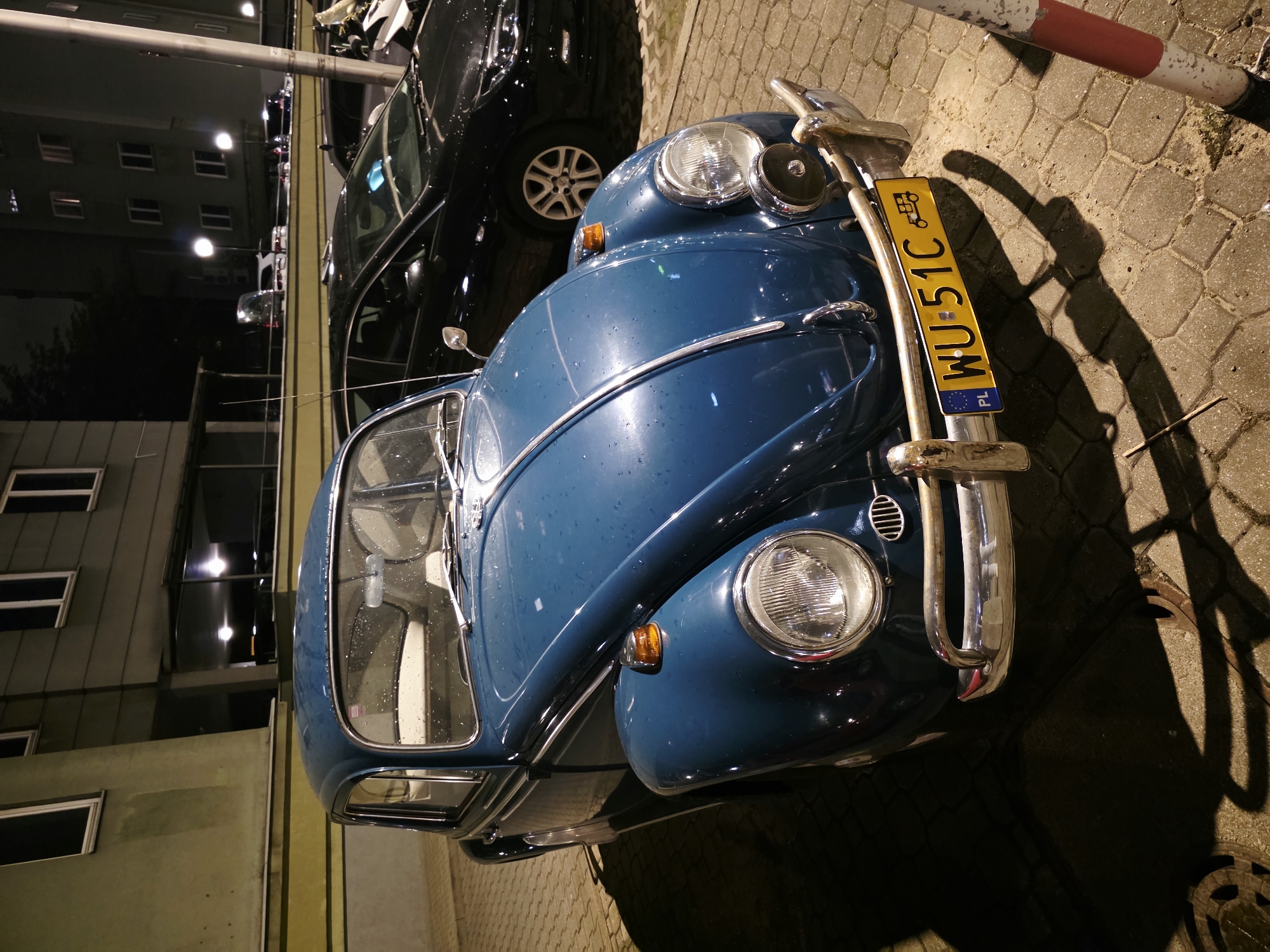 An eye-level medium shot captures a vintage blue Volkswagen Beetle parked on a cobblestone street at night. The car is angled slightly towards the viewer, with its front end prominently displayed. The car's paint is wet, suggesting recent rain, and it reflects the ambient light from streetlamps. The license plate, VU 51C, is clearly visible, with a Polish flag emblem. Behind the Beetle, a black modern sedan is parked. The background features a building with multiple windows, some of which are illuminated. The scene appears to be a city street in Warsaw, Poland, indicated by the license plate and the architectural style of the building.