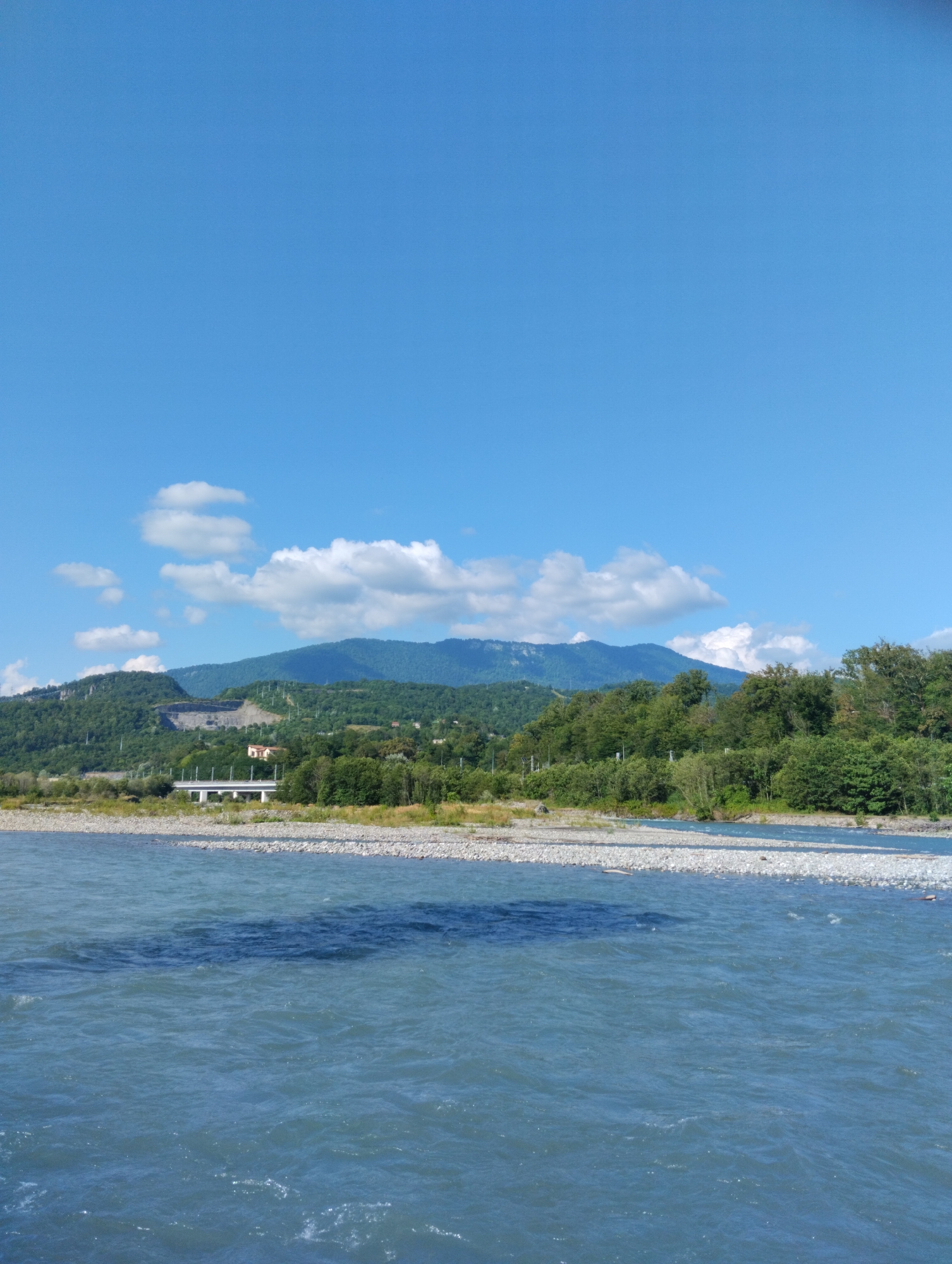 The image depicts a scenic view of a river flowing through a valley with lush green hills and mountains in the background. The sky is a clear blue with a few scattered white clouds. A bridge can be seen crossing the river in the mid-ground, with trees and foliage lining the riverbanks. The water in the river is a clear blue, with ripples on its surface. The scene is tranquil and serene, suggesting a natural and undeveloped environment.