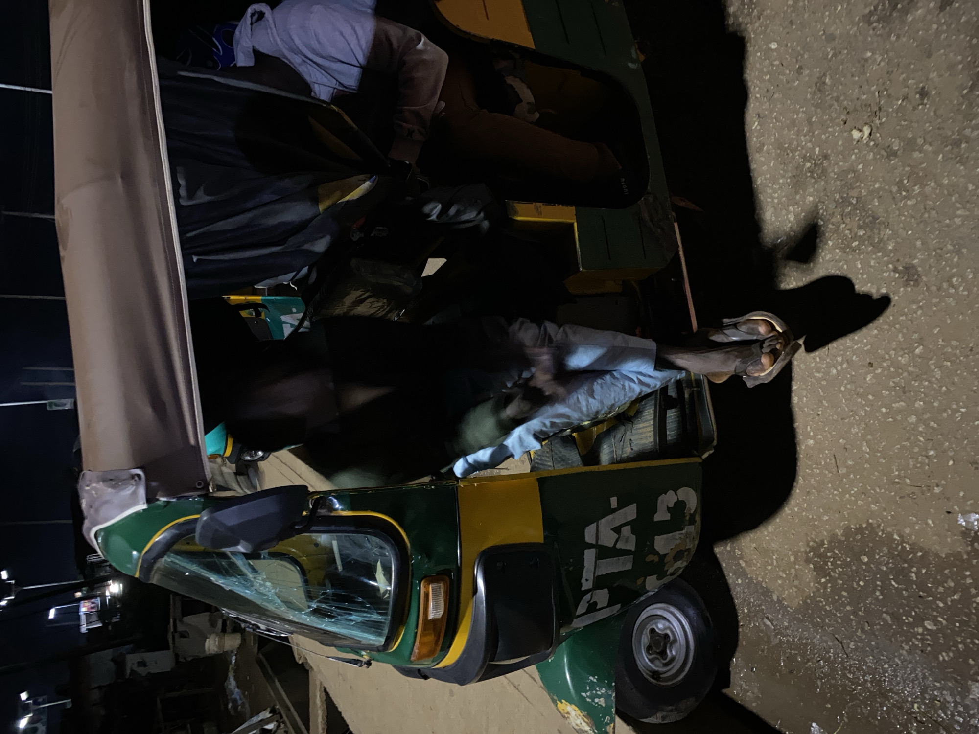 An auto-rickshaw, painted green and yellow with the partial text "PTA-13" on its side, is parked on a wet, asphalt surface in an urban environment at night. The windshield of the auto-rickshaw is cracked and broken. Inside the vehicle, three individuals are visible. One person, wearing a white hoodie and brown pants, is seated in the passenger area. Another person, wearing light blue pants, has their legs extended out of the vehicle, with their feet resting on the ground. A third person, largely obscured by a dark tarp or cloth, is also within the auto-rickshaw. The background suggests a commercial or industrial area with streetlights and structures visible in the distance. The overall scene appears to be in Jos, Nigeria, given the context provided.