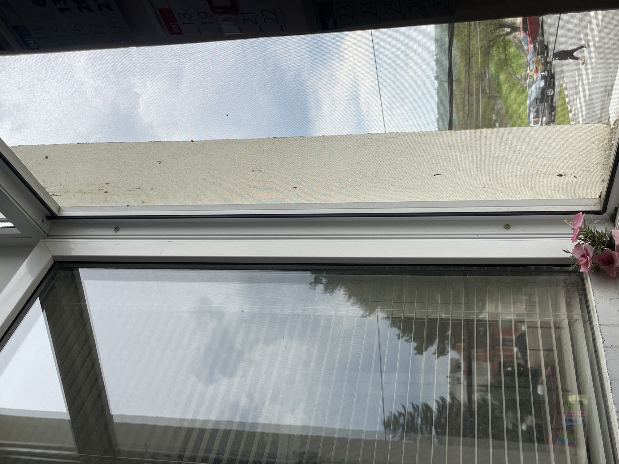 The image depicts a view through a window, obscured by a screen, with a calendar partially visible at the top. The window frame is white, and the scene outside includes a street with vehicles and pedestrians. The calendar shows dates ranging from April 14, 2026 to June 26, 2026. The image does not contain any specific information about the location in Sungurlare, Bulgaria.